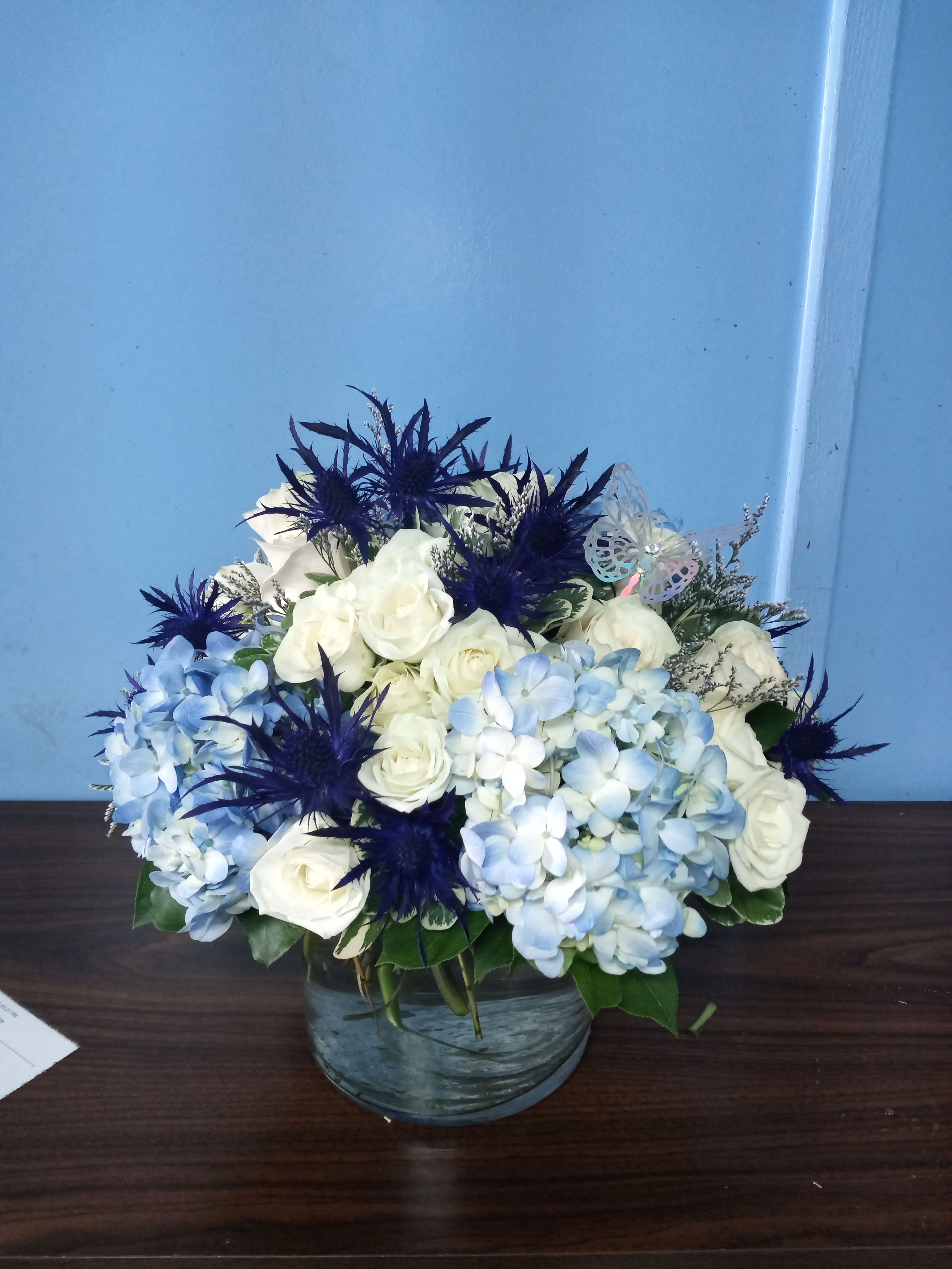 Thistle Whistle - Roundy moundy arrangement with blue hydrangeas, white roses and thistle in a 5 by 5 cylinder. One of our favorite designs.
