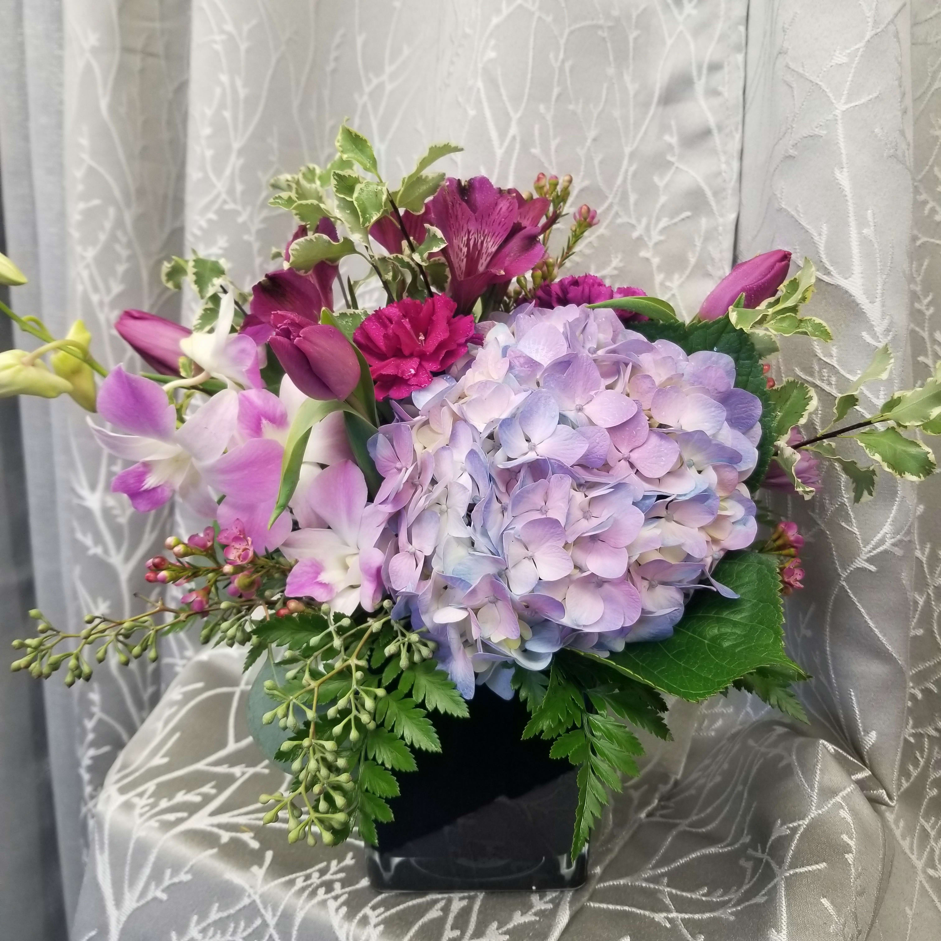 Love Spell by MG Florist  - Designed in a glass cube with beautiful shades of violet flowers. Starting with a hydrangea, tulips, dendrobium orchid,  alstromeria and accented with mini carnations.