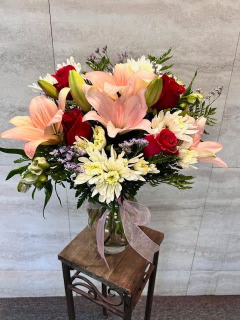 Spring Bouquet - This is a graceful mixed bouquet arranged in a clear glass vase. It features soft pink lilies as the focal flowers, giving a fresh, just-blooming feel. Deep red roses are tucked throughout, adding richness and contrast to the pastel tones.  The bouquet is complemented by white chrysanthemum-style blooms that bring fullness and texture, along with delicate lavender filler flowers and airy greenery that soften the overall look. A pale pink ribbon tied around the vase adds a gentle, elegant finishing touch.  Overall, the arrangement feels romantic, balanced, and fresh, blending warm colors with light, natural greenery for a classic yet cheerful floral display. 