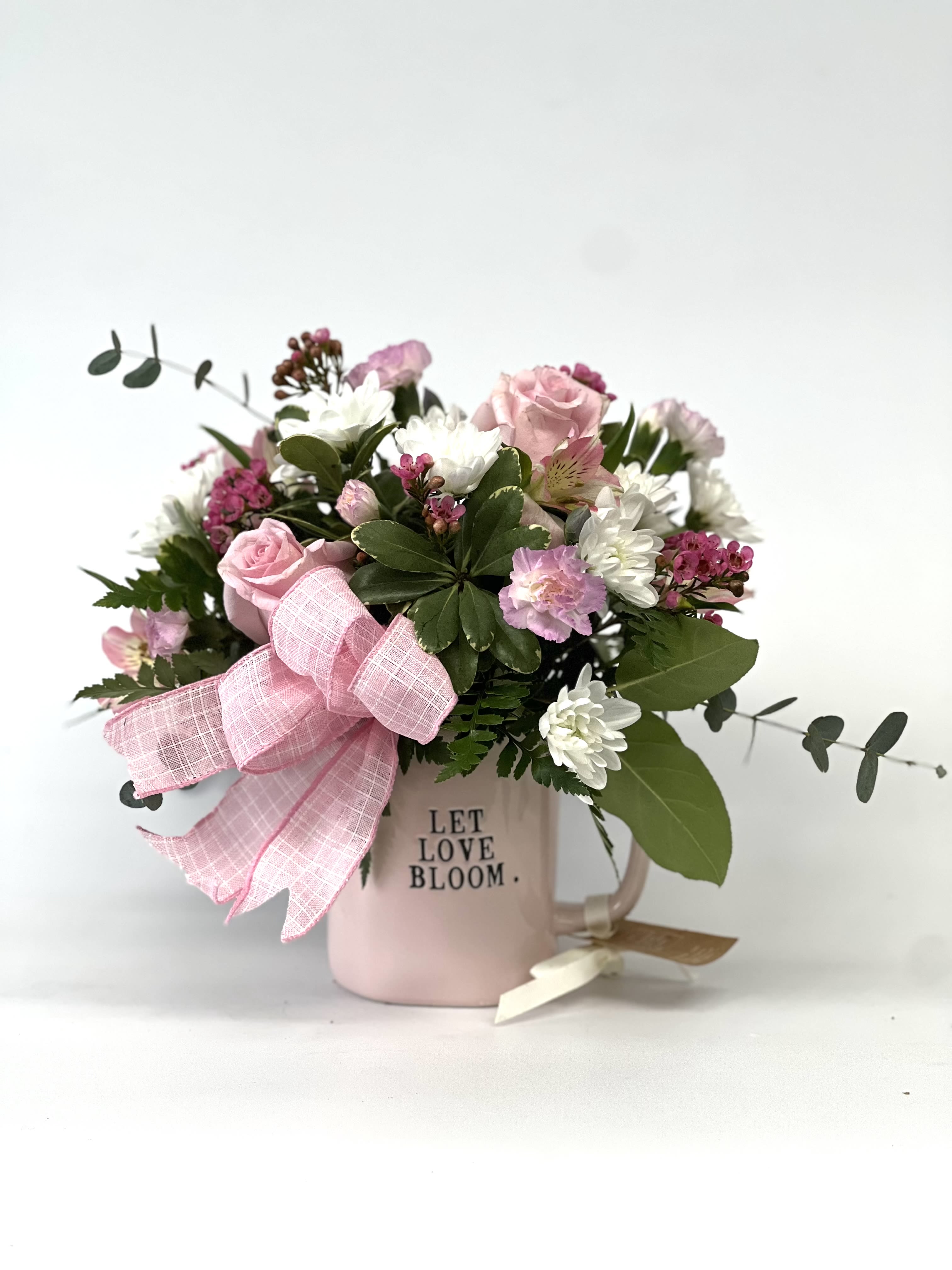 Bloom Mug Garden - The Bloom Mug Garden is a charming, heart-warming floral design that celebrates love, comfort, and simple beauty. This arrangement features a delicate mix of soft pink roses, fresh white blooms, textured seasonal flowers, and lush greenery, thoughtfully arranged in a keepsake ceramic mug and finished with a sweet coordinating ribbon.  With its gentle pastel tones and garden-inspired feel, this design is perfect for birthdays, thank-you gifts, encouragement, new beginnings, or just because. Designed to bring a smile and brighten any space, it’s as meaningful as it is beautiful.  Color Palette:  Soft Pink |  White |  Fresh Green  Please note: Flower varieties, colors, ribbon styles, and mug/vase design may vary due to seasonal and local availability. Each arrangement is uniquely crafted while maintaining the same overall look, feel, and color harmony.