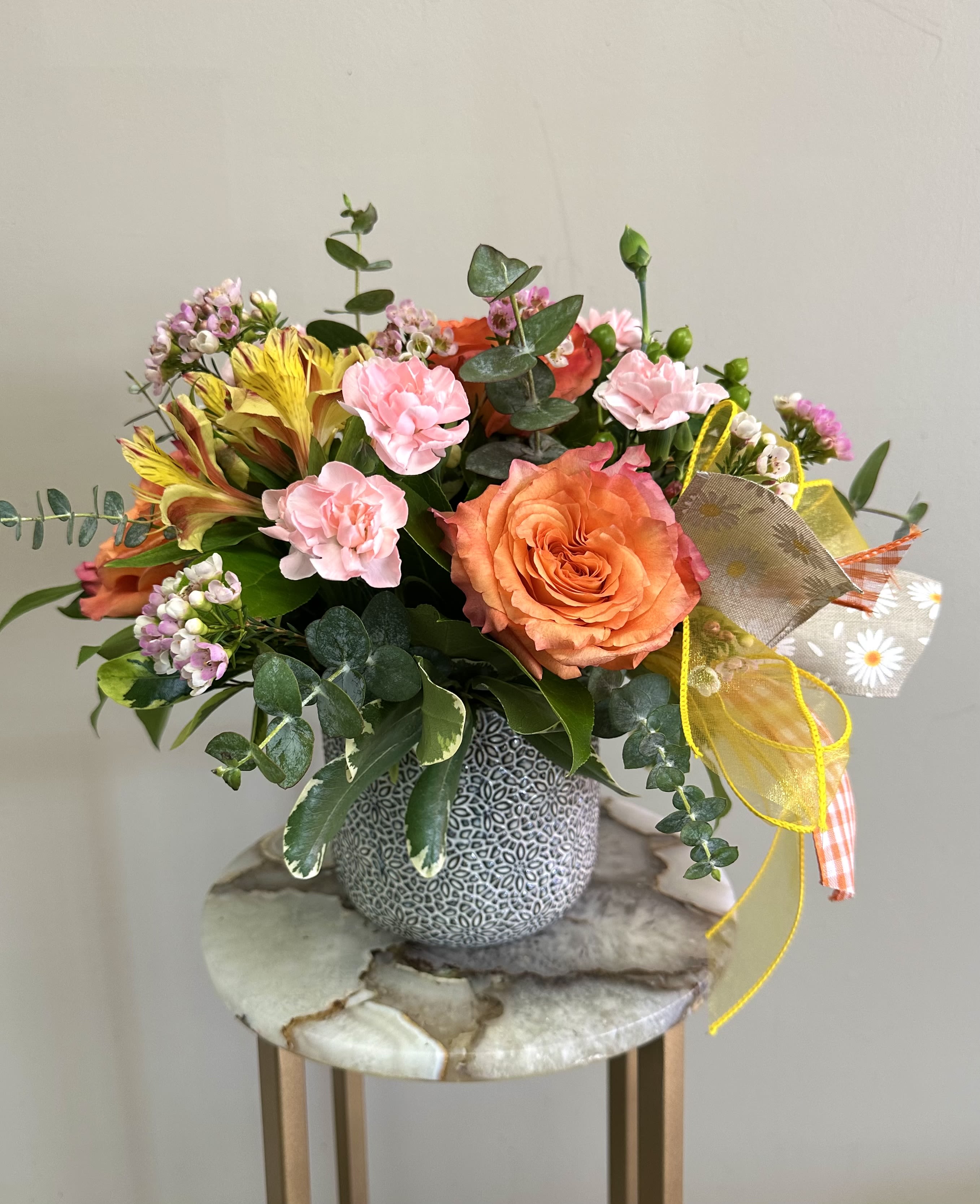 Sweet Meadow Garden - Soft and charming, Sweet Garden Meadow is a delightful mix of orange roses, yellow alstroemeria, light pink mini carnations, green hypericum, and pink wax flower, accented with spiral eucalyptus for a fresh, garden-inspired feel. Designed in a white ceramic pot with a textured floral design, this arrangement adds a touch of rustic elegance to any space.  Perfect for brightening someone's day or celebrating a special moment! 