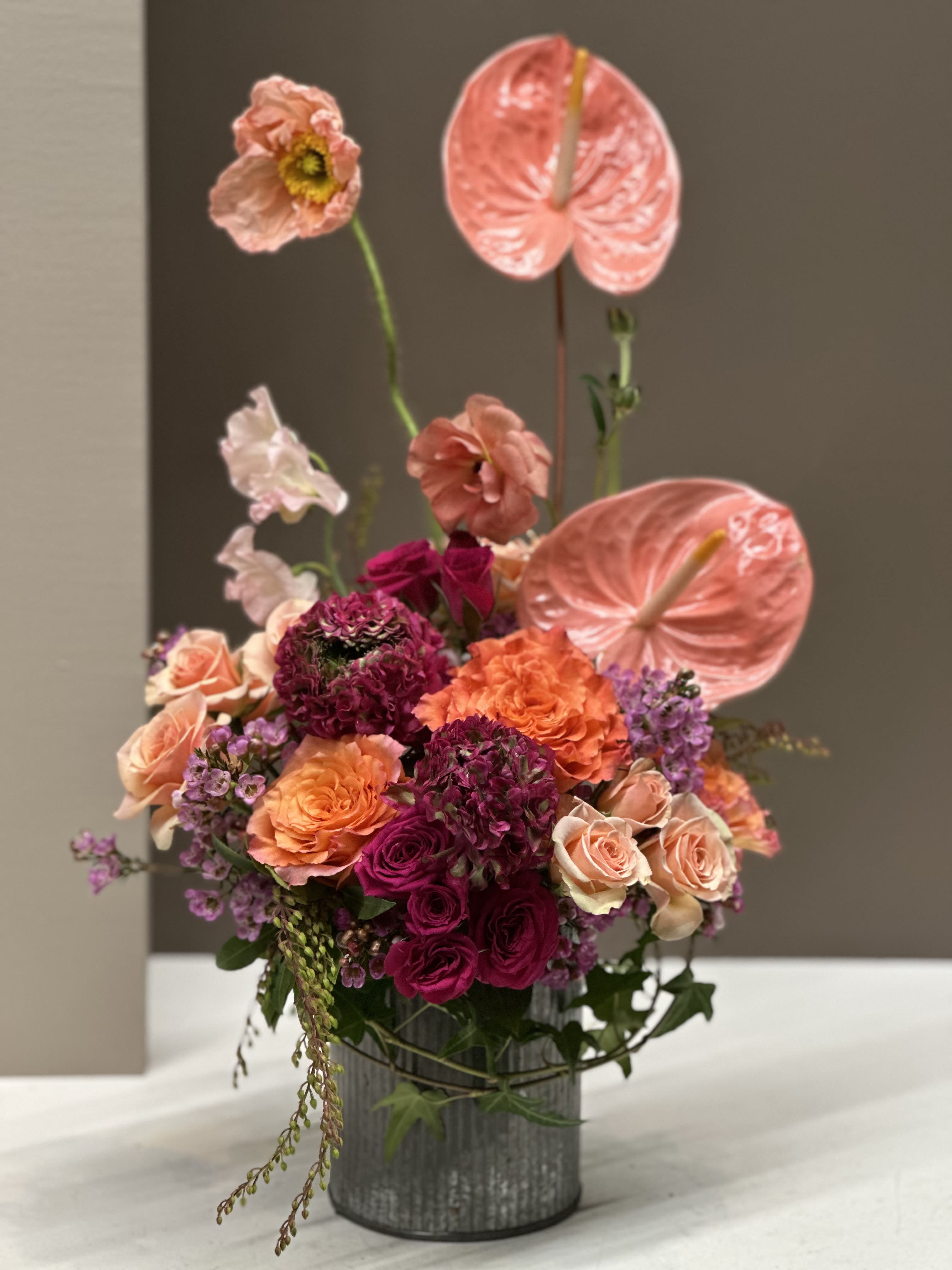 Peach Horizon - A striking modern arrangement in glorious sunset tones featuring Peach Anthurium; spray Roses in Peach, Coral, and Magenta; Mauve variegated Ranunculus; and accented with delicate Blush Waxflower. Statuesque stems of Anthurium and Poppy rise above the composition, creating dynamic height and a contemporary sculptural feel. Housed in a sleek galvanized metal vase, this bouquet blends soft, romantic tones with modern edge, making it a standout.