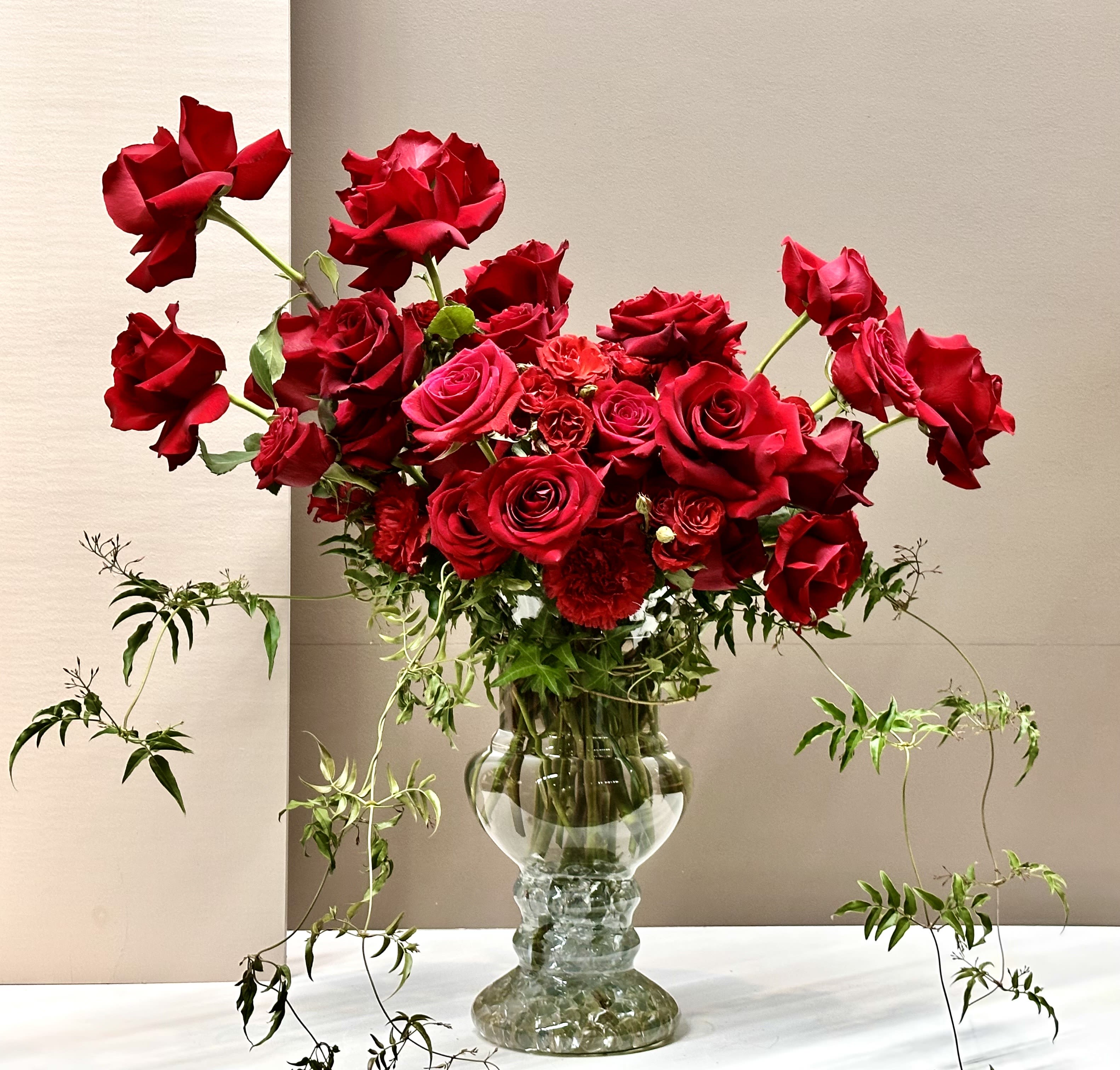 Scarlet Heights - Overflowing with elegance and drama, this taller arrangement features an abundant collection of Red Roses in varied sizes, thoughtfully arranged to create a sense of lavish fullness. Some blooms reach skyward, adding movement and height, while delicate trailing foliage cascades gracefully, softening the structure and lending a natural charm. Housed in a substantial glass vase, it exudes romance.