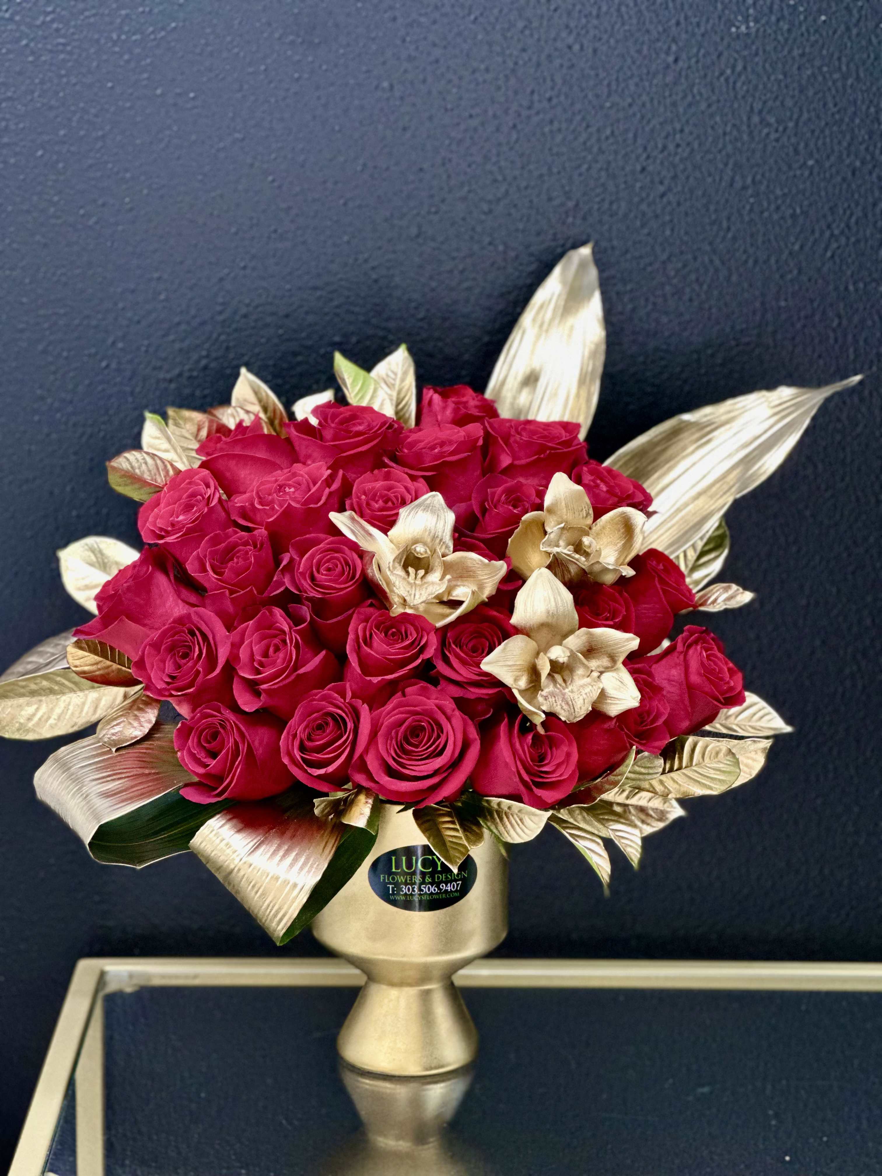 The Golden Touch  - The Golden Touch blends the richness of deep red roses with shimmering gold accents for a truly captivating design. Gold-sprayed foliage and orchids add a layer of sophistication, all set in a luxurious gold ceramic vessel that elevates the arrangement into a timeless centerpiece. Perfect for anniversaries, romantic gestures, and unforgettable celebrations.  Arrangement Includes: -Fresh Cut Roses, Orchids and Other Blooms -Gold Ceramic Vase -Care Instructions -Custom Greeting Card