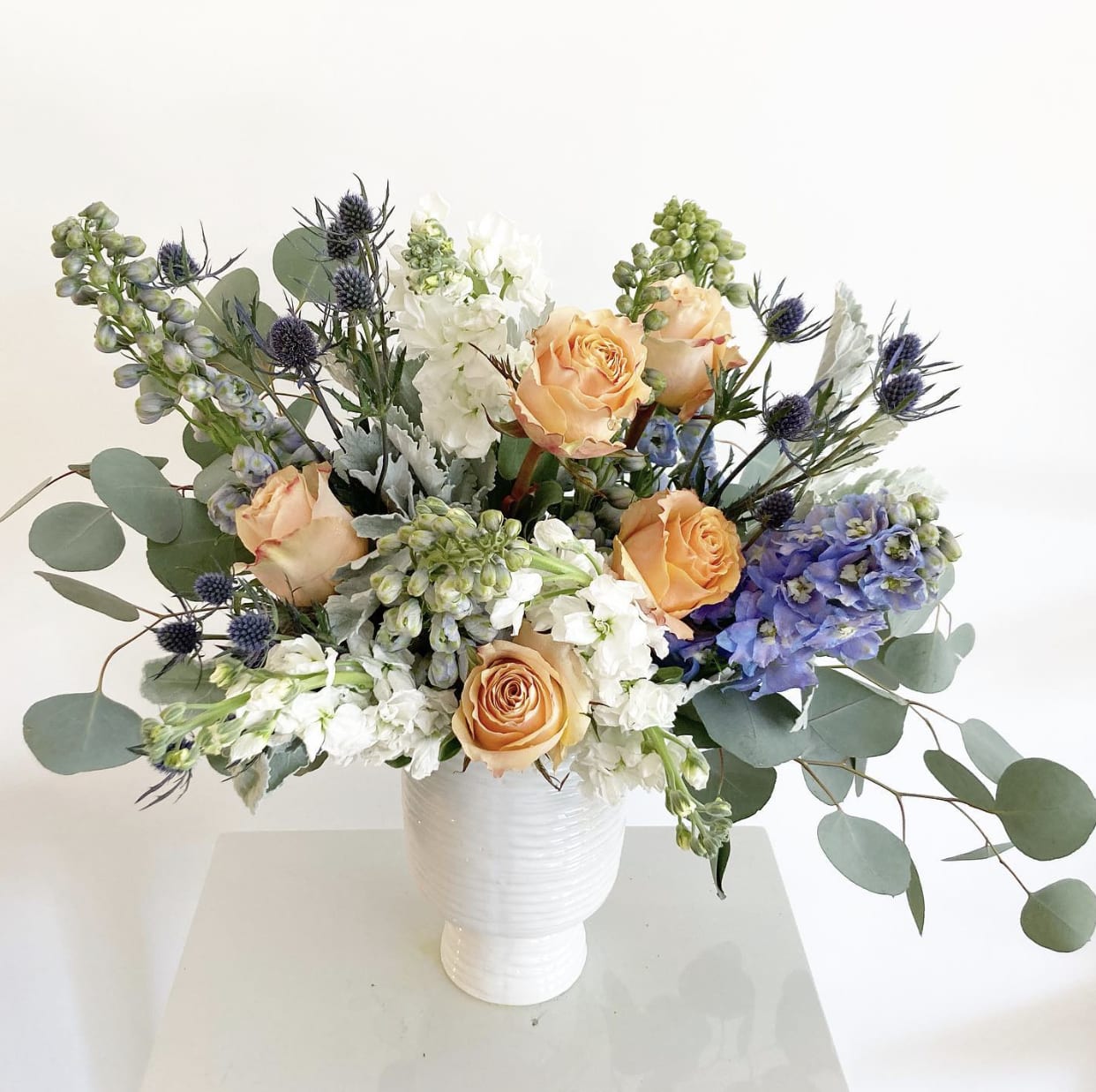 The Cora - The Cora is a captivating arrangement that balances elegance with whimsy. Soft, glowing peach shimmer roses take center stage, complemented by delicate white stock, airy light blue delphinium, and striking accents of dark blue thistle. Silvery touches of dusty miller and silver dollar eucalyptus add sophistication, all beautifully arranged in a sleek white ceramic container.