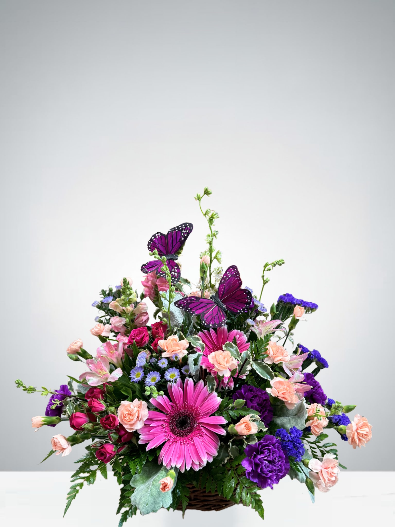 Butterfly Dreams - Delicate and whimsical, this charming arrangement features pink gerbers, purple carnations, soft peach mini carnation, hot pink spray roses, pink snapdragons, alstroemeria and cheerful statice, all nestled among lush greenery. Two graceful faux butterflies rest gently atop the blooms, adding a magical touch. Designed in a woven basket, it’s perfect for birthdays, get-well wishes, or simply bringing a smile to someone’s face. 