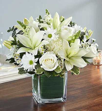 cube arranement - Arrangement of white roses, Asiatic lilies, football mums, snapdragon, daisy poms and monte casino; accented with fresh greenery Artistically designed by our florists in a classic clear glass cube lined with a Ti leaf ribbon; vase measures 5"H x 5"D Large arrangement measures approximately 11"H x 11"L Medium arrangement measures approximately 10"H x 10"L Small arrangement does not include roses and measures approximately 9"H x 9"L Perfect gift for delivery to the homes of family members or friends Our florists hand-design each arrangement, so colors, varieties, and container may vary due to local availability Lilies may arrive in bud form and will open to full beauty over the next 2-3 days