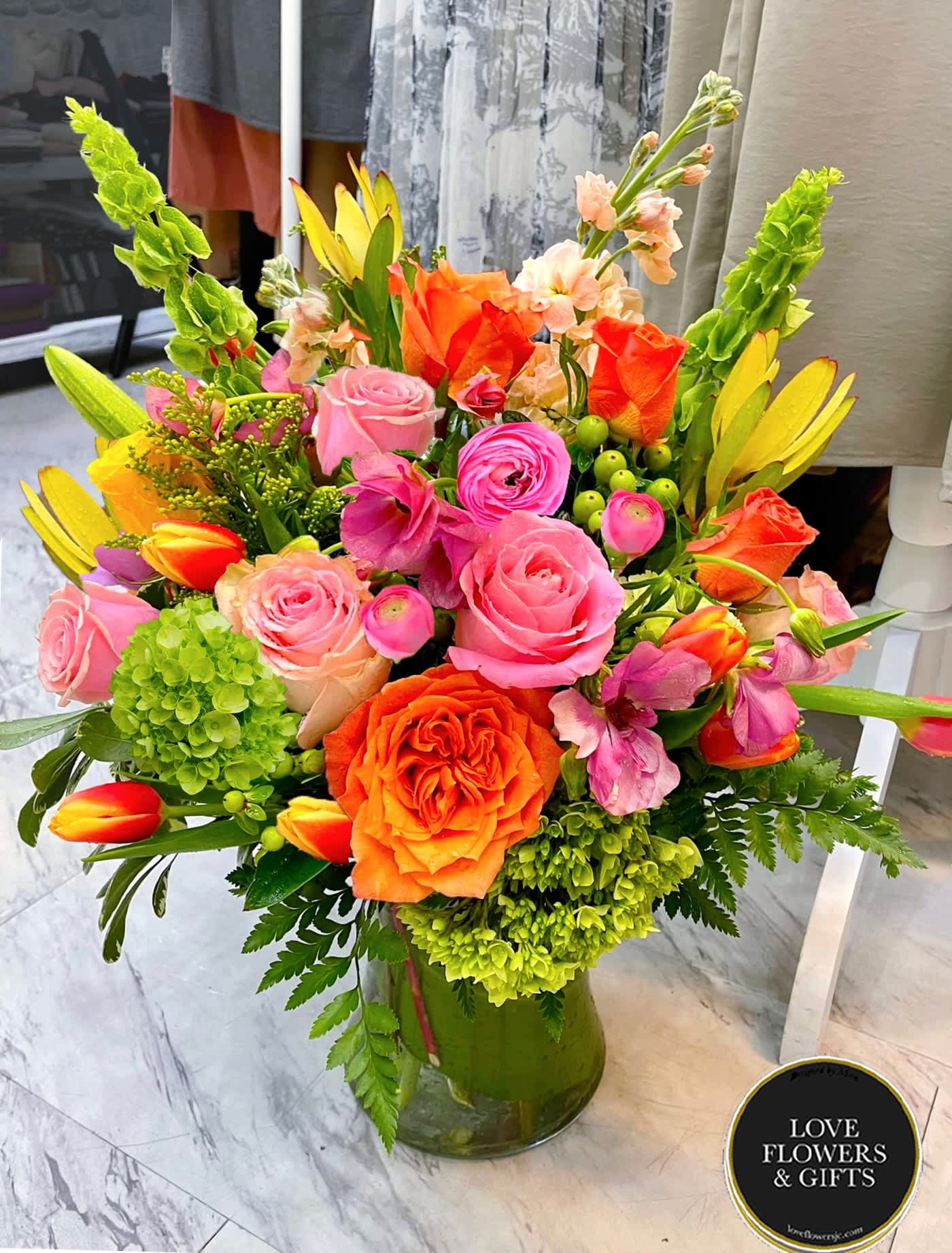 Prima Gala  - A luxurious and abundant spring arrangement made of ranunculus, roses, tulips, snapdragons, and more fresh flowers in a glass vase wrapped in banana leaves.  View from all angles, 360 Degrees. 