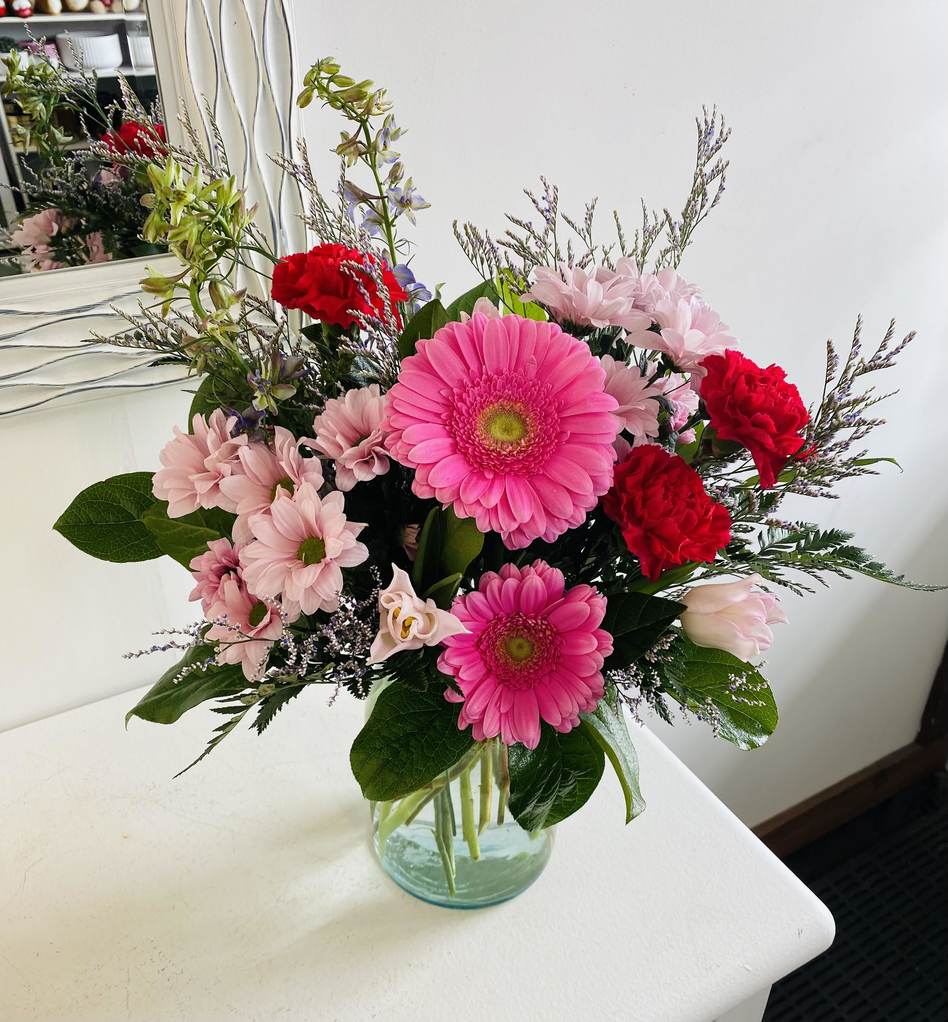 Raspberry Punch - Bursting with cheer, this vibrant arrangement pairs bold hot pink gerberas with playful red carnations and soft pink daisies for a joyful display of color. Wisps of lavender-toned filler and rich greenery add texture and movement, all beautifully gathered in a clear glass vase. It's the perfect bouquet to brighten someone’s day or add a pop of fun to any space.