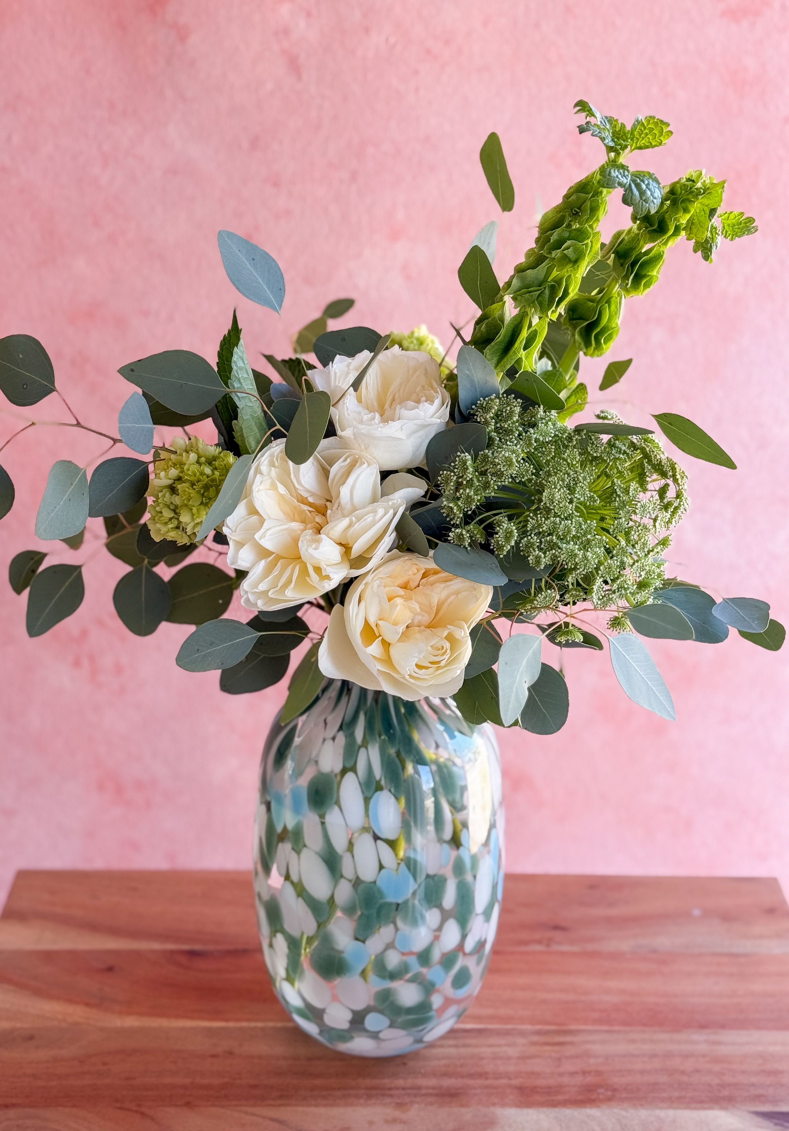 Zia  - A delicate arrangement with lush greenery and white flowers in a glass artisan vase. 