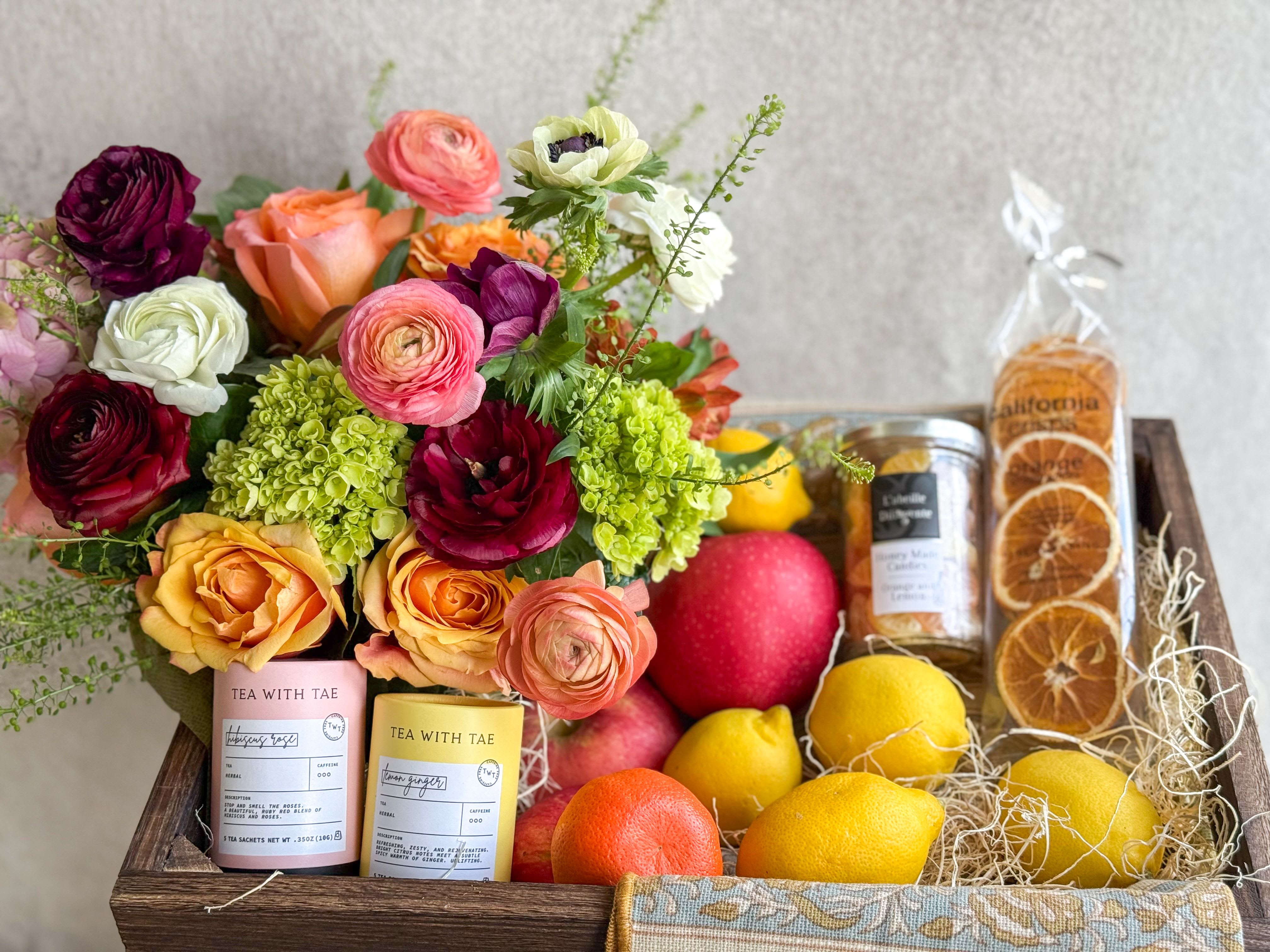 Flowers, Fruit &amp; Tea Basket - Gift in good taste with this elegant assortment of treats. A seasonal lush floral arrangement, floral pattern tea towel, Assortment of fresh fruit, lemon and hibiscus tea, French Orange Lemon Honey Candy, and dried orange slice snacks. All designed in a large wooden crate, includes handwritten card.   You deserve the best. If an item is unavailable, we’ll sub in products of equal or greater value. 