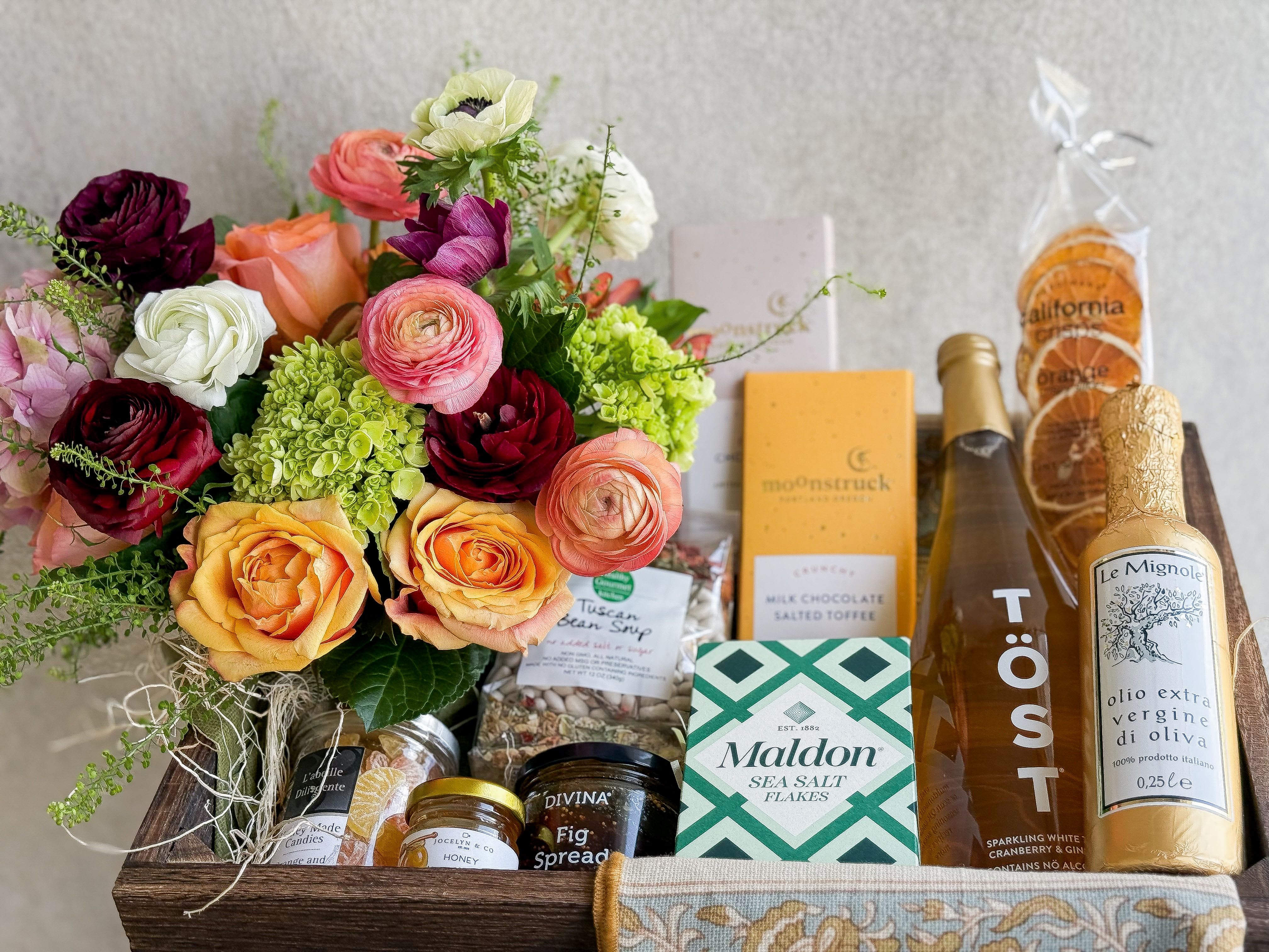 Gourmet Gift Basket - Gift in good taste with this gourmet gift basket. A seasonal lush floral arrangement, floral pattern tea towel, French Orange Lemon Honey Candy, and  extra virgin olive oil,  maldon sea salt, fig spread, tuscan bean soup, 2 chocolate bars, honey, and dried orange slice snacks. All designed in a large wooden crate, includes handwritten card.   You deserve the best. If an item is unavailable, we’ll sub in products of equal or greater value.