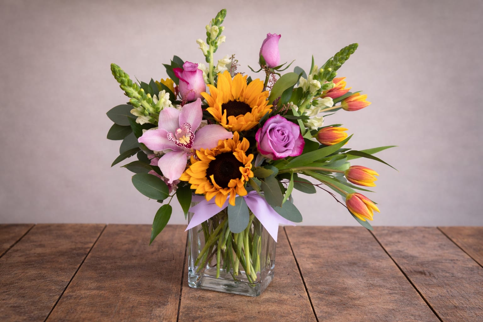 Sunny Wild  - Bursting with bright, carefree charm, the Sunny Wild arrangement blends the warmth of golden sunflowers with the soft elegance of lavender roses, pink tulips, and exotic orchids. Lush greenery and snapdragons add height and texture, creating a cheerful yet refined design that feels fresh from the garden. Perfect for celebrating special moments or simply spreading sunshine.  Please note that every design is custom made and substitution to container and flower types may happen from time to time. Design aspects will always stay the same, but if needed, we will substitute with flowers of equal or greater value to offer you a great floral shopping experience! Our flowers are 100% guaranteed. Please call us with any questions or concerns