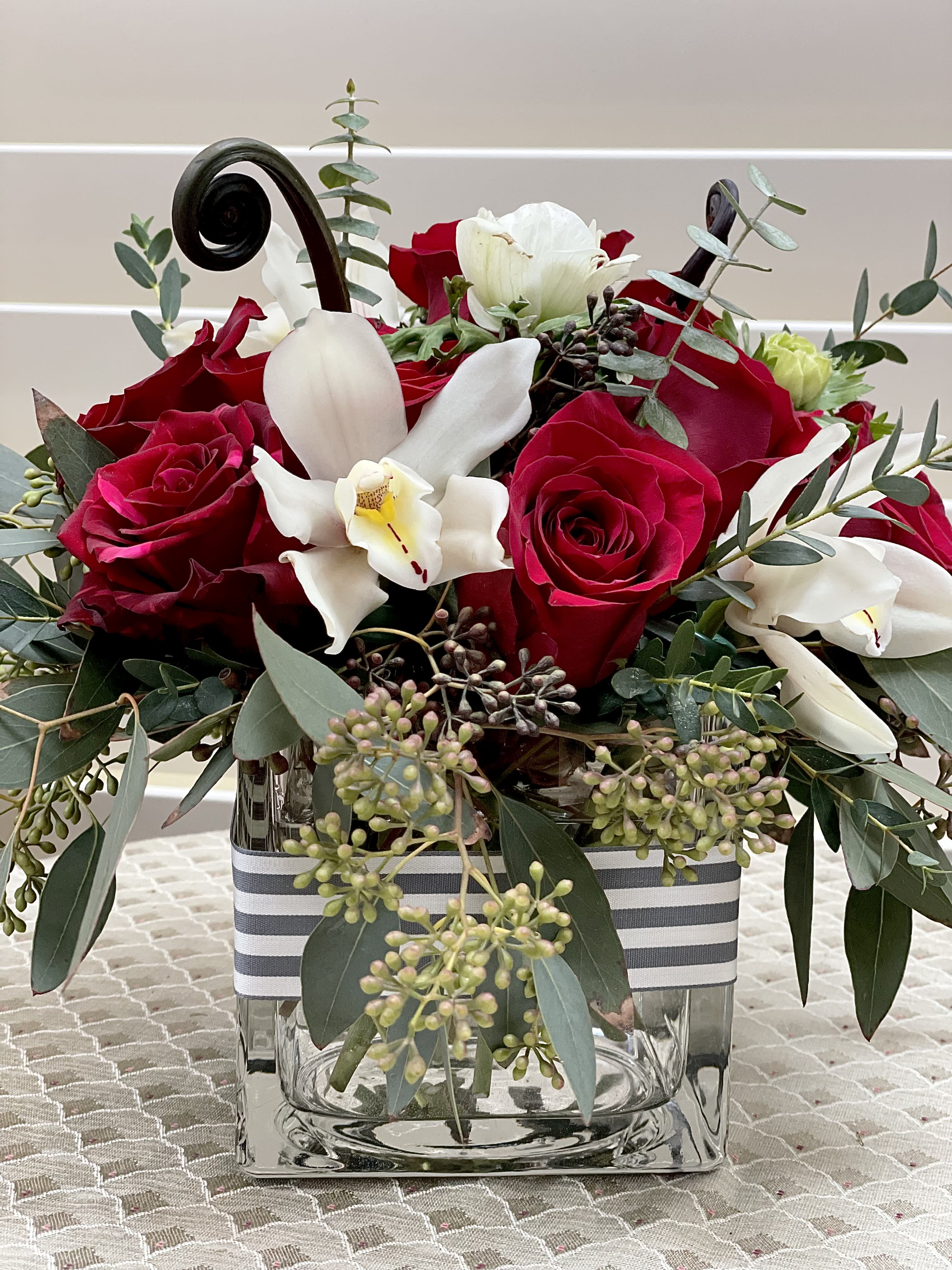 Orchid and Rose Delight - A design of red roses with orchids and other seasonal blooms, nestled against a variety of greens and textural elements.