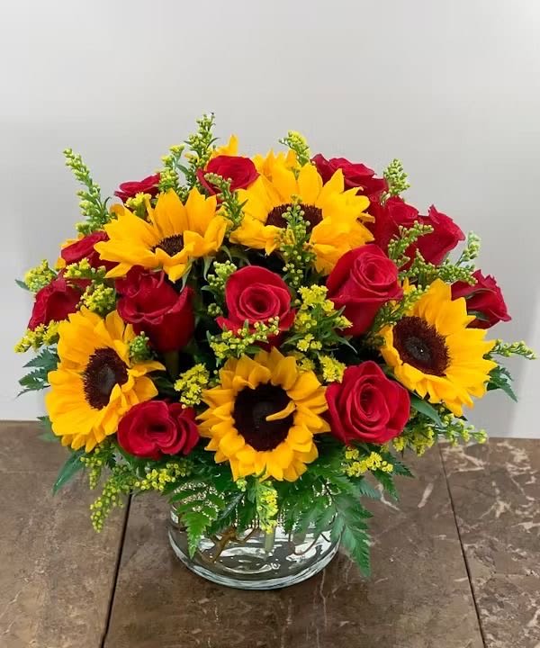 Roses &amp; Sunflowers - A bright and cheerful arrangement featuring vibrant sunflowers paired with classic red roses, accented with fresh greenery and golden filler blooms. Designed in a clear glass vase, this piece is perfect for celebrating love, happiness, and warm wishes—a bold pop of color for any occasion.