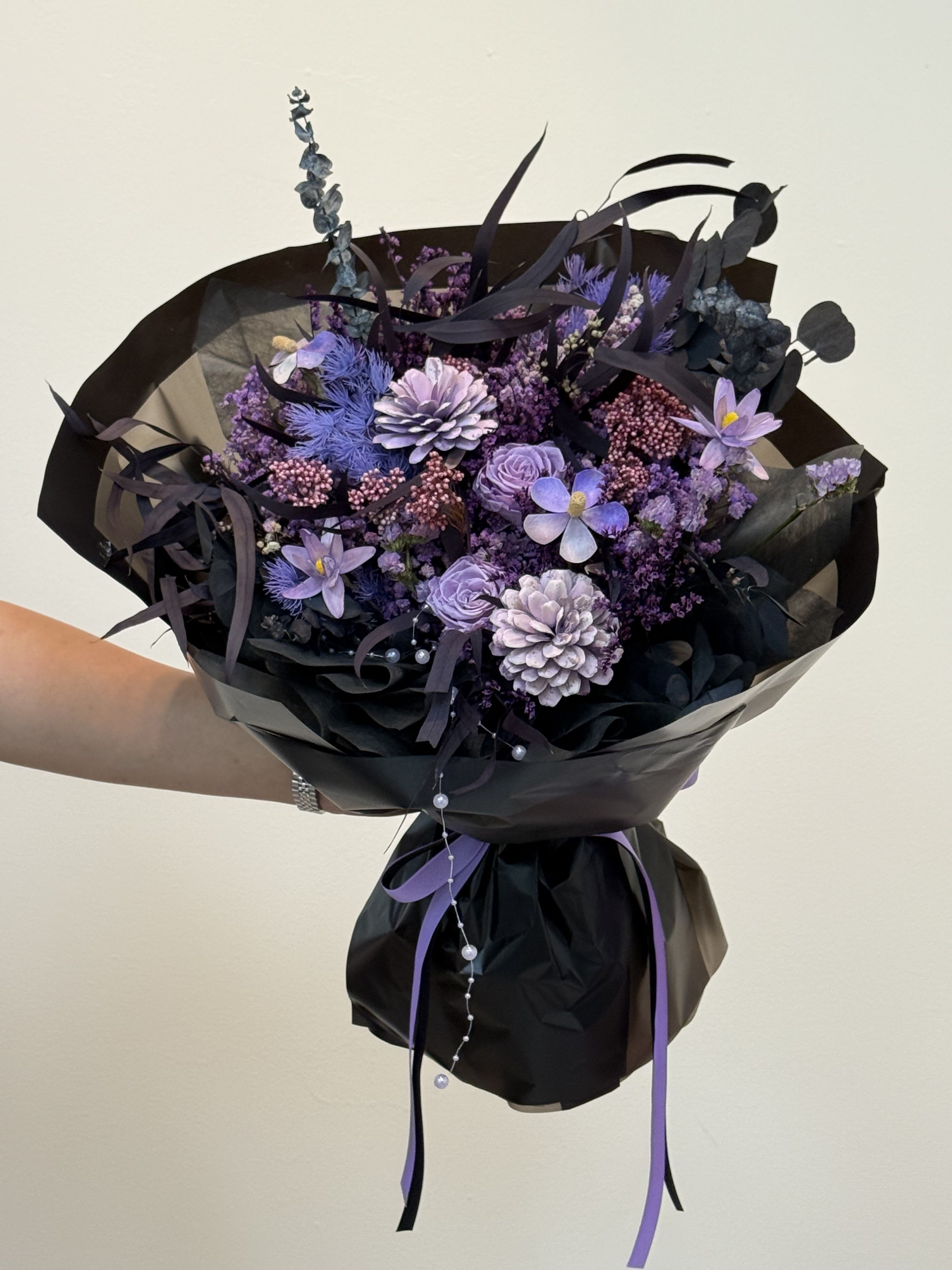 Silent Midnight - This is a mixed bouquet of dark purple and black, with a mysterious and elegant style. It is usually composed of a variety of flower materials, creating unique visual effects. Main flowers: mainly purple and near-black flowers of different shades. Common ones include dark purple or lavender roses, hydrangeas, oranges and tulips. White flowers (such as chrysanthemums or fresies) are used as decorations to provide contrast. Matching flowers and leaves: dark-toned filled flowers and dark green or blackened leaves such as eucalyptus leaves enhance the overall mysterious atmosphere. Packaging style The bouquet is wrapped in black wrapping paper, which highlights the color of the flower itself, creating a sense of drama and high-end. Finally, it was tied with a purple ribbon and decorated with some small beads to add a sense of exquisiteness. Symbolic meaning Purple usually symbolizes mystery, nobility, romance and elegance. The black packaging brings a sense of avant-garde, uniqueness and power, making this bouquet very suitable for birthdays, anniversaries or any occasions that require special and bold expressions.
