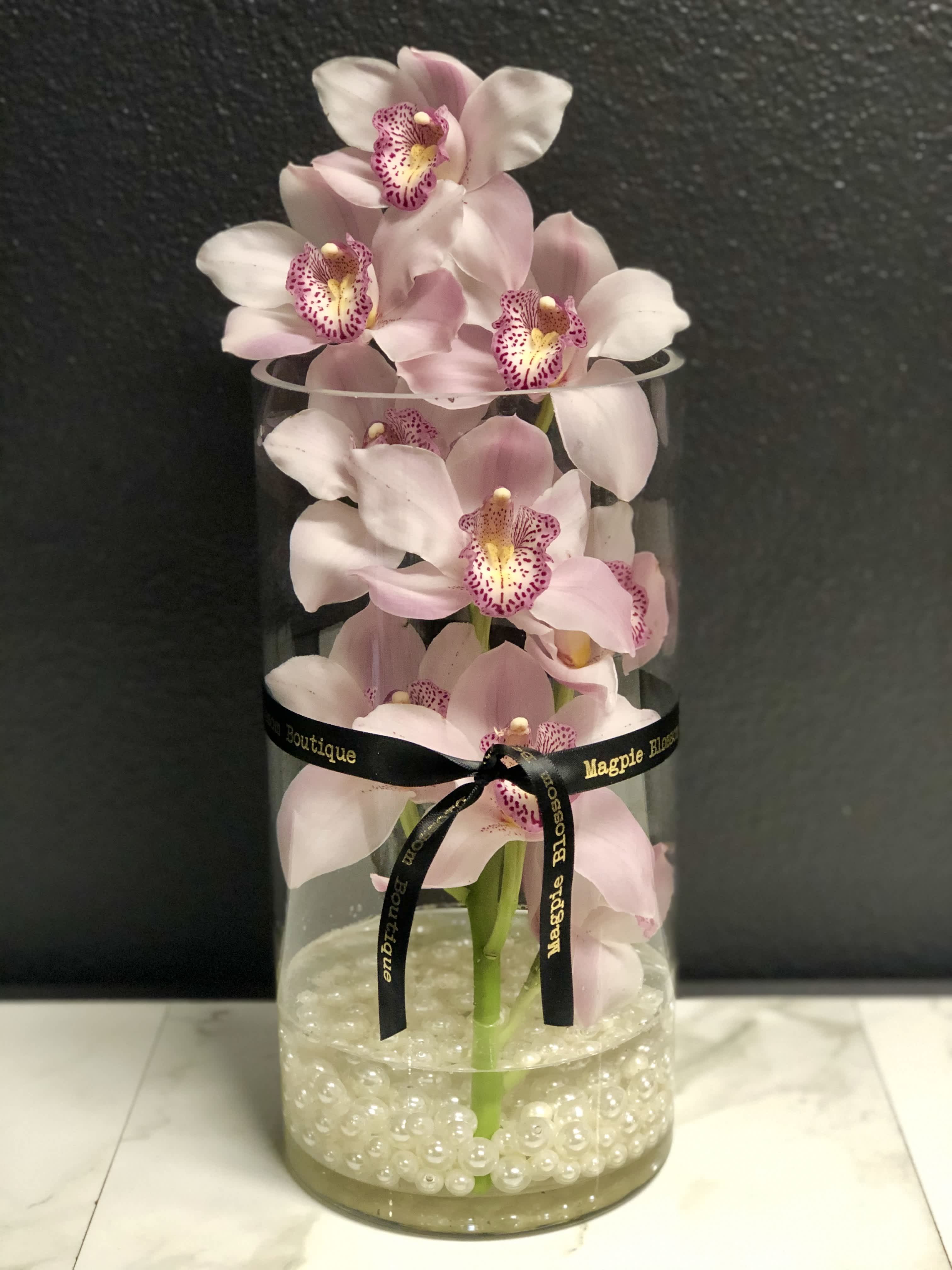 Valentine's Day Orchid Token - Available 2/11-2/14 Only - Sleek and Elegant. A fresh-cut Cymbidium Orchid stem in a clear glass vase with a scoop of feminine pearls.  Orchid color may vary.   Approx. 12+" tall. 