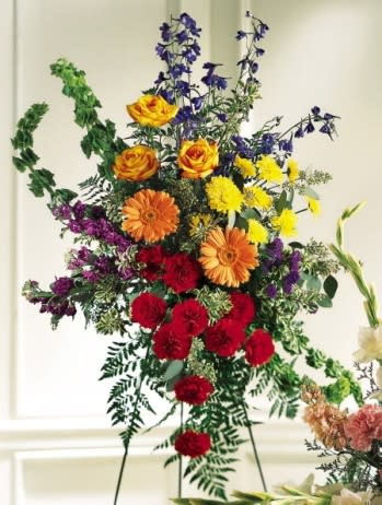 Fond Farewell Standing Spray - The colorful arrangement includes orange gerbera daisies, red carnations, yellow roses, blue belladonna and more. Delivered with an easel, this spray is appropriate to send to a funeral home or service