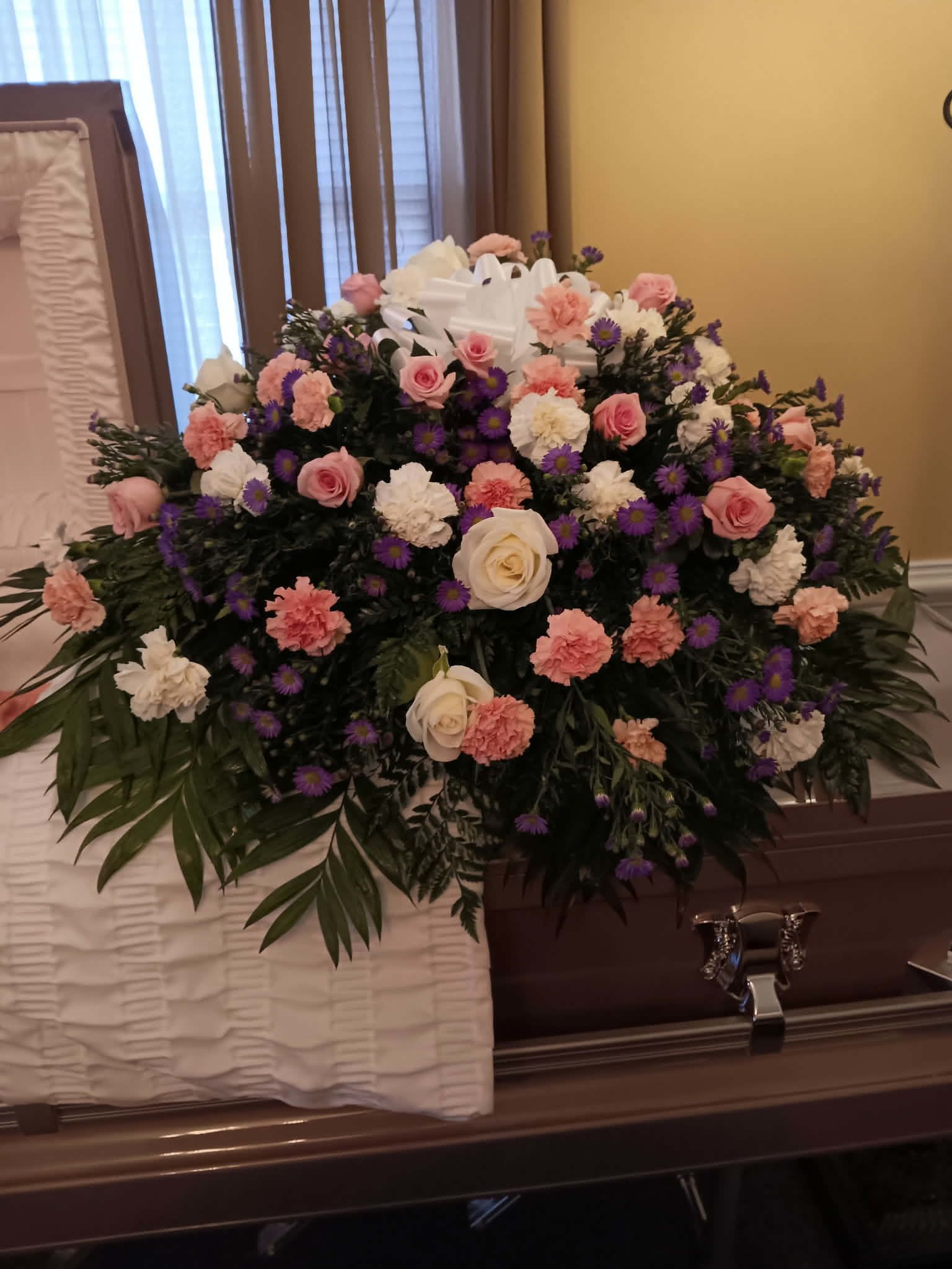 A Little Touch of Purple Casket Spray - A beautiful casket spray with white/pink roses, pink/white carnations, purple montecasino, with lush greenery to fill, and a bow.