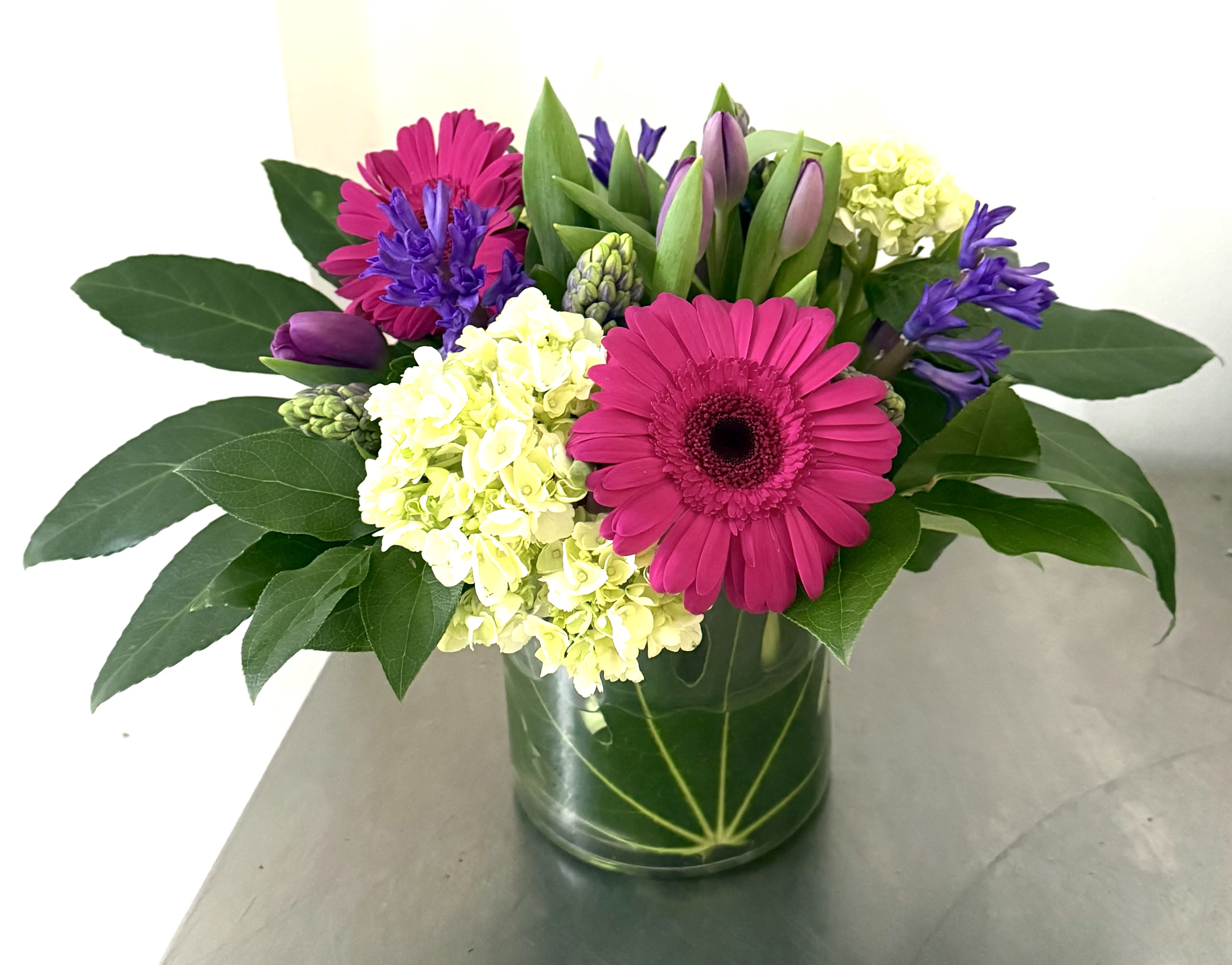 Vibrant Early Spring Garden - A stunning, garden-style arrangement presented in a clear glass cube vase, which is elegantly lined with a large green leaf. The design bursts with a vibrant mix of hot pink gerbera daisies, lime green hydrangeas, purple tulips, and other complementary purple and hyacinths. This cheerful assortment brings the bright and bold shades of spring indoors. 