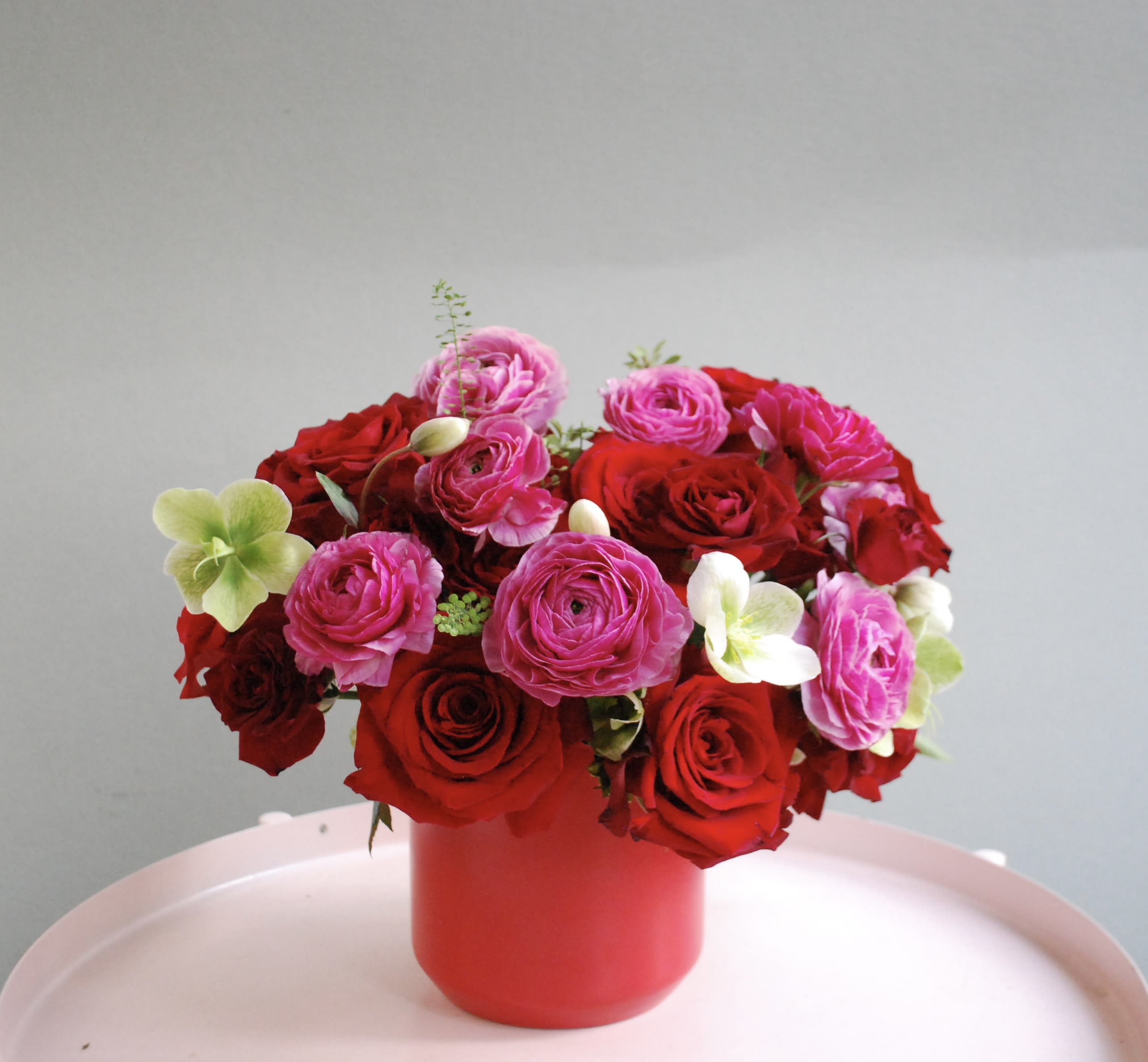 I’ts you - A bold and romantic arrangement featuring lush red roses paired with vibrant pink garden roses and ranunculus. Soft accents of pale green hellebore and delicate seasonal greens add contrast and movement, creating a modern yet timeless design. Arranged in a sleek red cylinder vase, this piece is perfect for anniversaries, Valentine’s Day, or any moment that calls for dramatic romance.