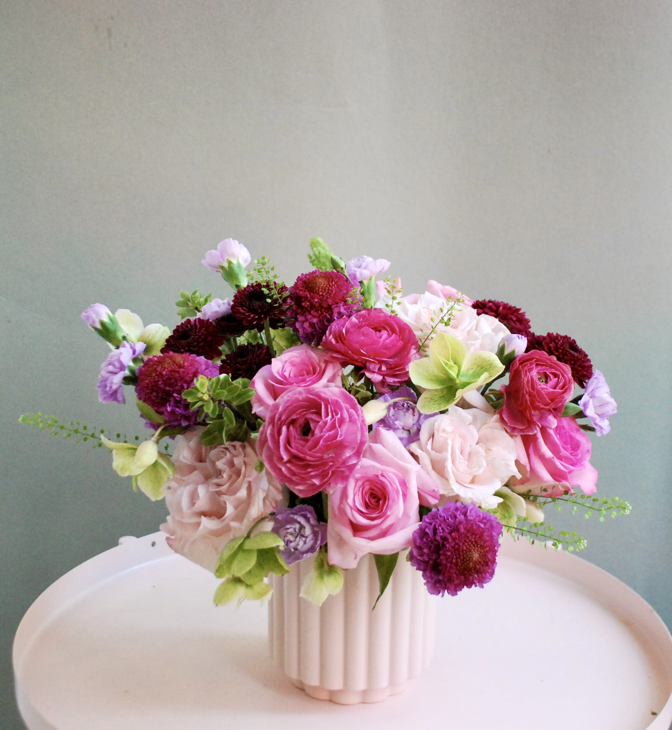 Cupid  - This lush, garden-inspired arrangement blends soft blush and hot pink roses with rich berry-toned blooms and playful seasonal accents. Textural layers of ranunculus, spray roses, and delicate greenery give this design a full, abundant feel. Styled in a white ribbed ceramic vase, it’s romantic, feminine, and effortlessly elegant—ideal for birthdays, celebrations, or just because.