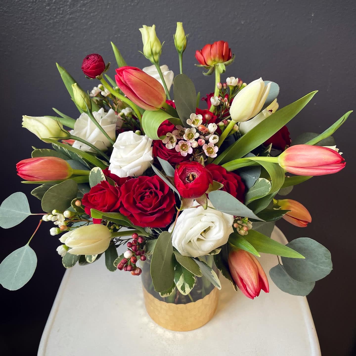 XOXO - Tulips + red spray roses + red ranunculus + white lisianthus arranged in a gold footed glass cylinder vase.  Approximately 13" tall x 12" wide 