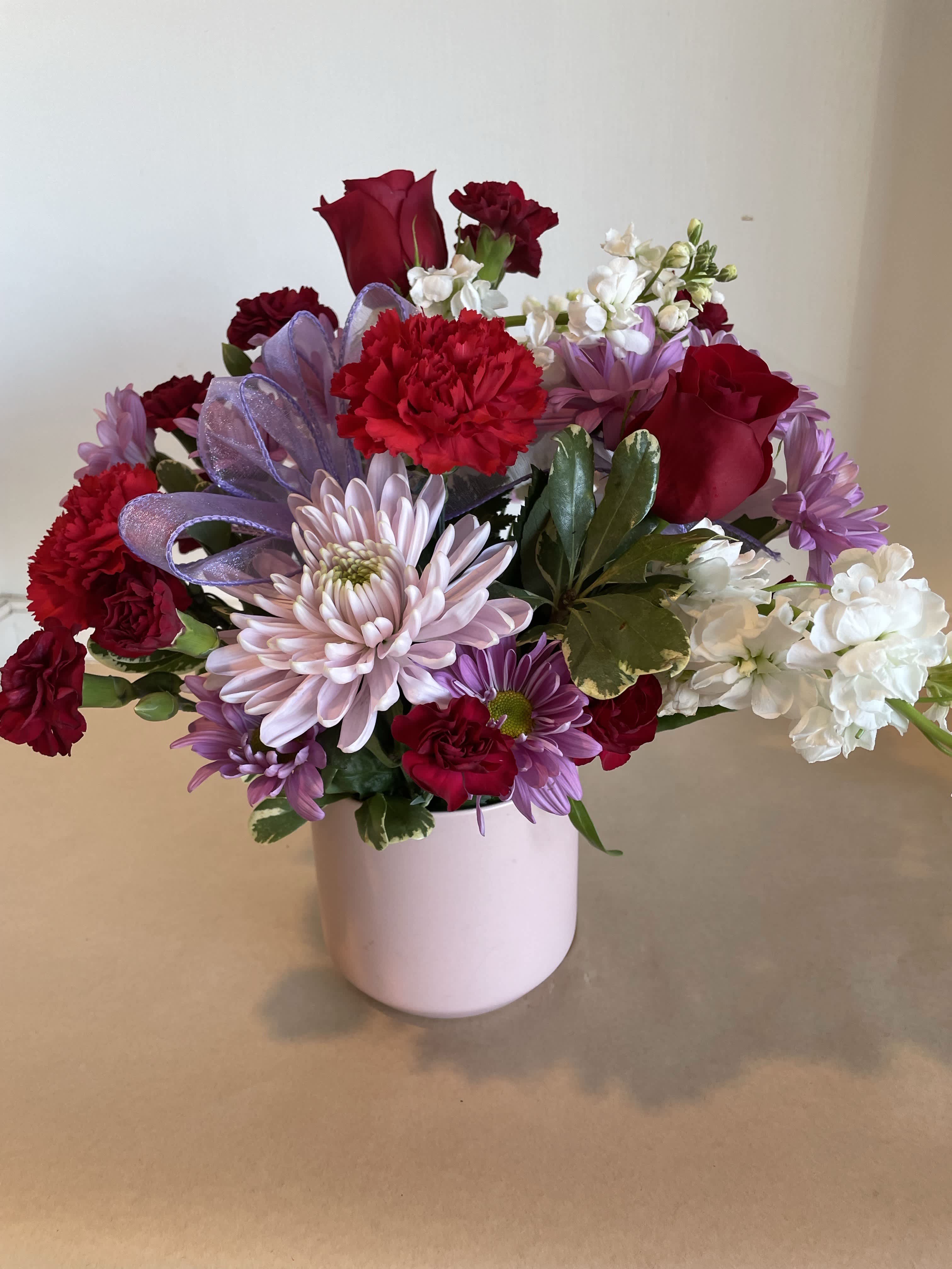 Love Story Bouquet - Show them you love them with our Love Story Bouquet. Arranged in a blush pink ceramic pot, we arrange our freshest blooms in shades of red, lavendars, and white. This arrangement includes a mix of daisies, roses, stock, and carnations.
