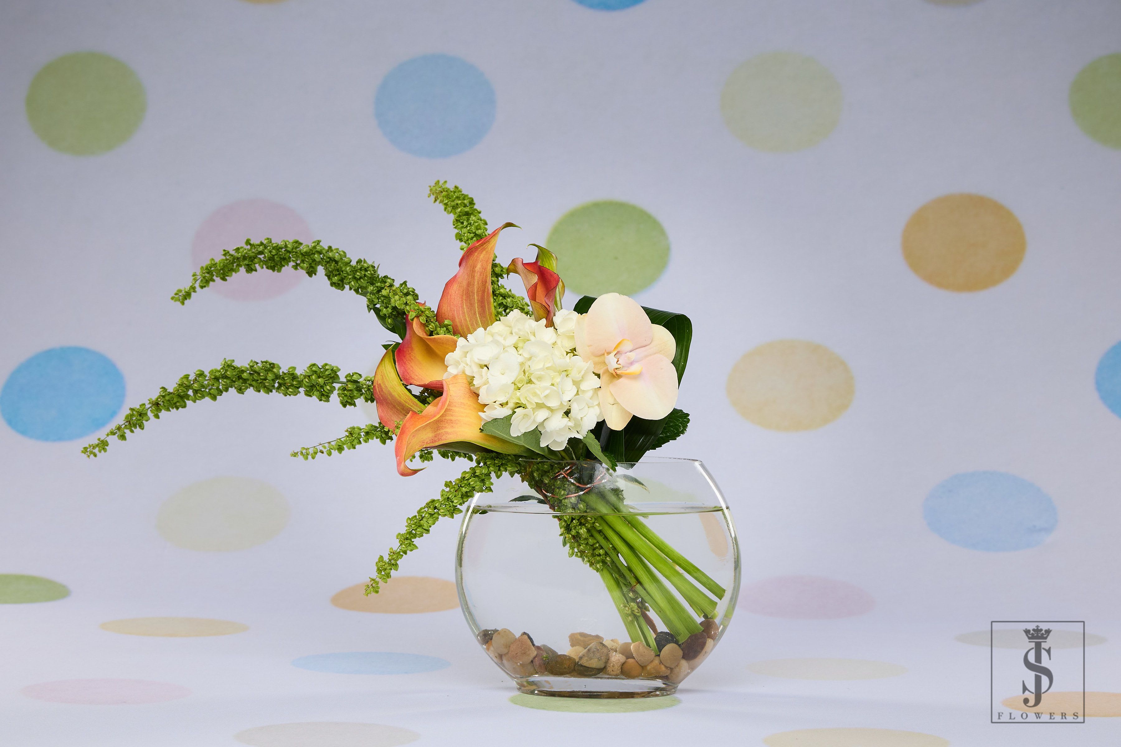 Whispering Elegance (Colors will vary) - Whispering Elegance is a serene and graceful floral arrangement that speaks in soft tones and subtle textures. A beautiful blend of peach calla lilies, creamy white hydrangeas, and a delicate orchid bloom form the centerpiece of this design. Flowing green amaranthus and rich aspidistra leaves add movement and modern flair. Set in a clear glass bowl vase with natural stones, this composition brings sophistication and balance into any space.  Perfect for sending love, celebrating achievements, or elevating modern interiors with quiet beauty.  