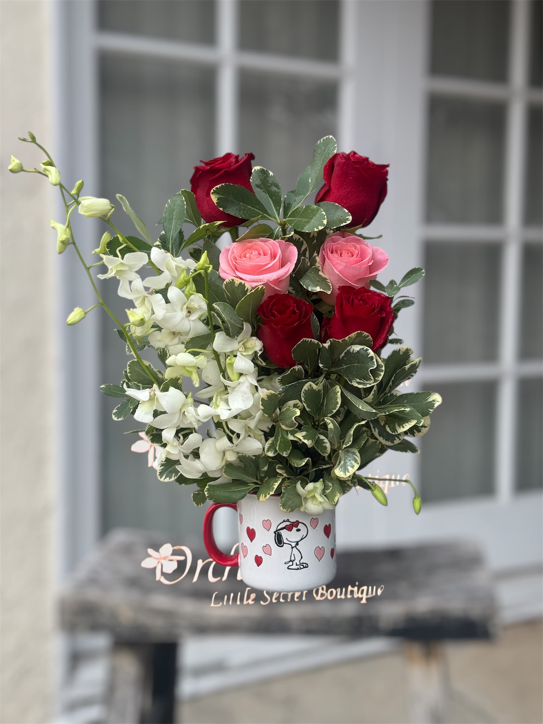 Red Pink Roses Orchids Sweetheart Snoopy  Mug Arrangement - Arrangement measures approximately 19”H -22"H Surprise a loved one with this charming and expressive floral arrangement, hand-crafted in a delightful, reusable Snoopy mug. This vibrant design combines the deep romance of red roses with the soft, blushing warmth of pink roses, as well as white exotic dendrobium orchids and variegated green foliage. The flowers are arranged in a white mug with a red handle, decorated with small red hearts and a drawing of Snoopy, creating a truly eye-catching gift for any occasion. Perfect for anniversaries, Valentine's Day, or simply to bring a smile, this arrangement is sure to make a lasting impression!     