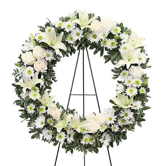 Purity Wreath - A classic all-white sympathy wreath designed with elegance and serenity. This wreath features graceful white lilies, white roses, and white daisies, accented with fresh green centers and lush foliage. The circular shape symbolizes eternity and everlasting remembrance, while the pure white palette represents peace, honor, and eternal rest. Displayed on an easel, this wreath is a timeless and dignified tribute to express love, respect, and heartfelt condolences.24 Inches Finish
