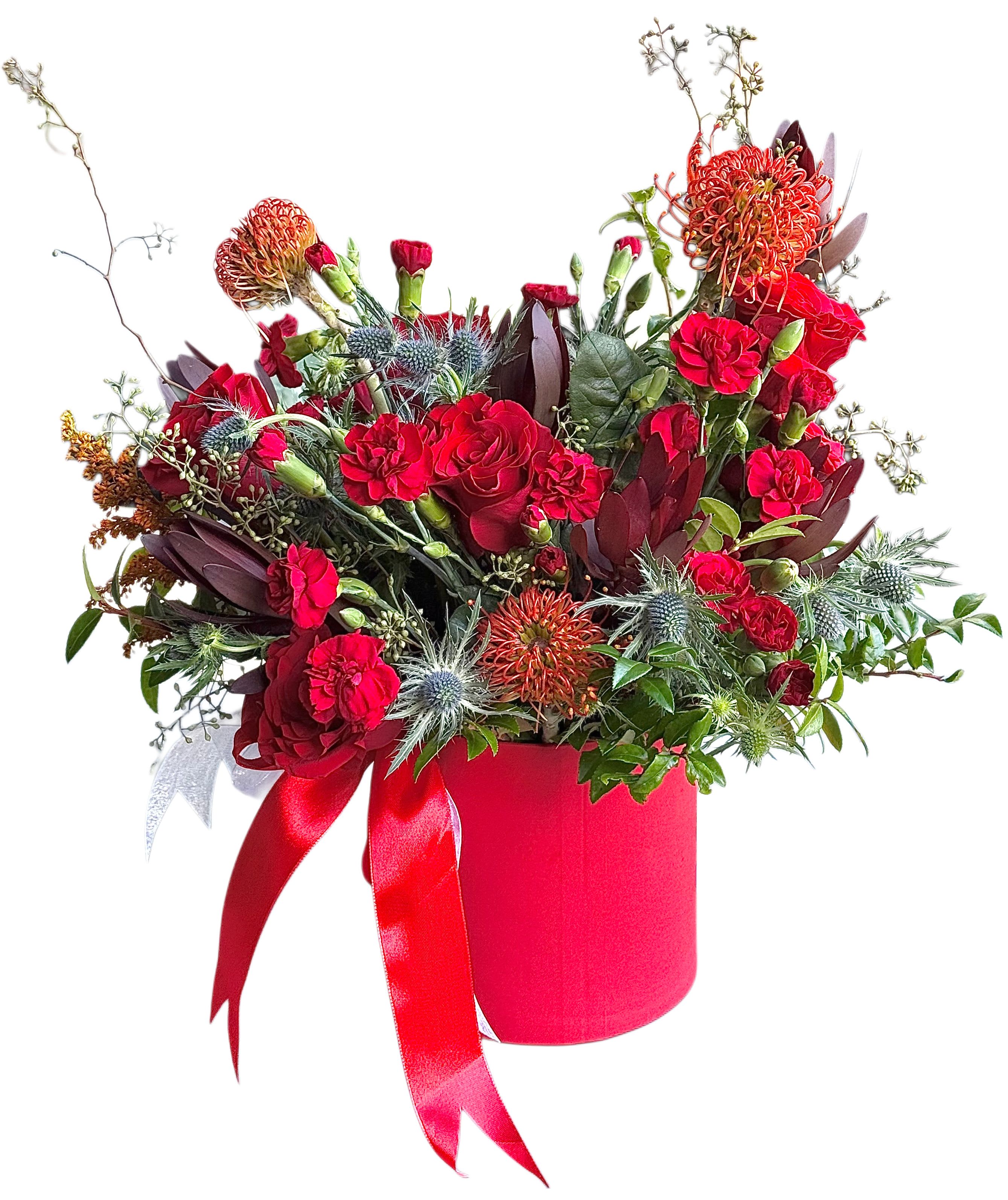 Crimson Devotion | Signature Red Box Arrangement - A bold and luxurious red floral arrangement designed to make a statement. This handcrafted hat box features premium red roses and carnations, accented with textured eryngium, seasonal blooms, and rich burgundy foliage. Arranged in a classic red velvet box and finished with elegant ribbon details, this piece embodies passion, strength, and timeless beauty.  Perfect for anniversaries, birthdays, romantic occasions, or moments when words are not enough. Each arrangement is uniquely styled while maintaining the same color palette and premium quality.