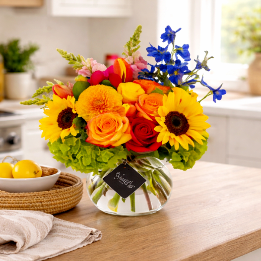 Midsummer Bloom - Bring joy and warmth into any space with our Colorburst Bloom arrangement — a radiant mix of sunflowers, roses, dahlias, tulips, snapdragons, delphiniums, and hydrangeas. Handcrafted in a clear glass vase, this vibrant bouquet features bold yellows, fiery oranges, soft pinks, and brilliant blues for a cheerful display that captures the essence of celebration. Perfect for celebrations, get-well wishes, or brightening someone's day, this floral arrangement is an unforgettable statement of love and happiness. Ideal for home decor or as a thoughtful gift,