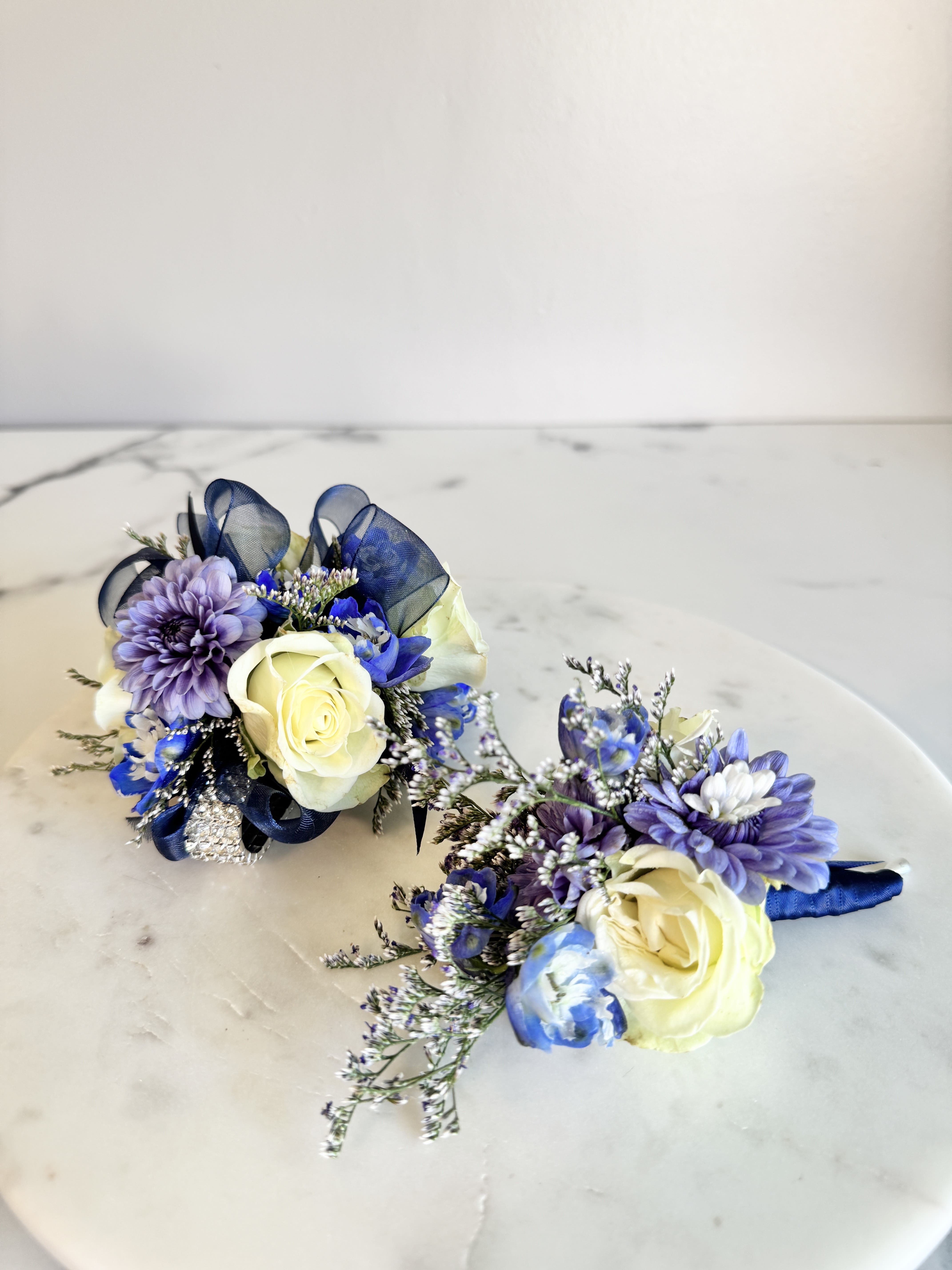 Corsage and Boutonniere Set - A youthful and elegant design especially created by the Florist. Flowers are hand selected by the Florist. Perfect for prom, homecoming, and school dance.  Please specify preferred color theme in the Florist Instructions when ordering. Colors can be in White, Pink, Blue, Lavender, Orange, Red, Yellow, Green or No Preference.  Corsages come with 2 strings of pearls OR on elegant elastic rhinestone band for DELUXE.  We highly advise ordering at least 2 days in advance to ensure availability of supplies for the Color Selection.   If your event falls on a weekend, choose Friday delivery. Corsage and Boutonniere will stay fresh for a few days if kept in a fridge. Delivered in a waterproof box with clear plastic lid.