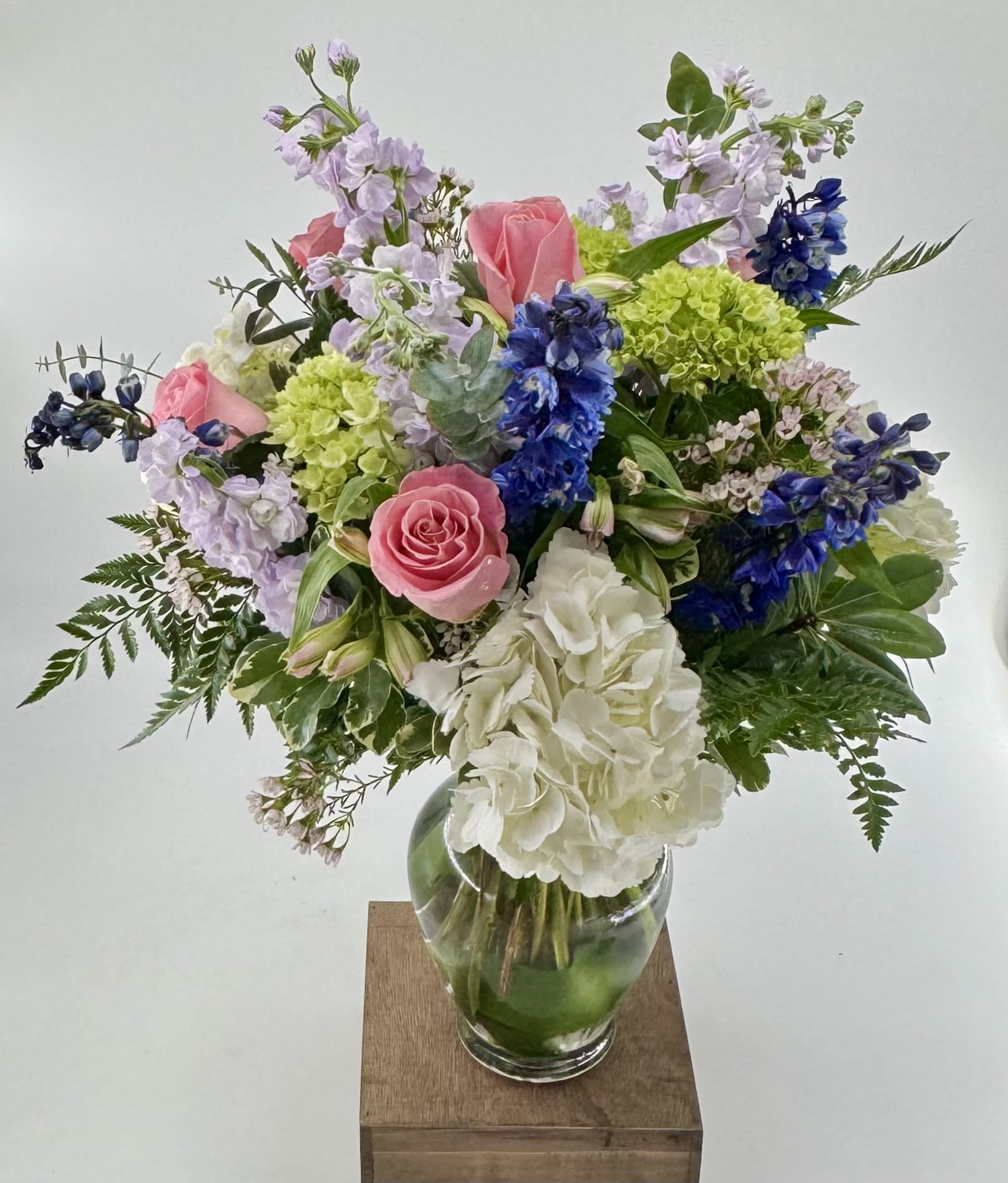Tranquil Oasis (DL) - Step into a whimsical realm with our "Tranquil Oasis" floral arrangement, a magical creation inspired by the allure of a mystical garden in full bloom. This enchanting ensemble features a captivating combination of delicate pink roses, lush green and white hydrangea, fragrant purple stock, and graceful blue delphinium.