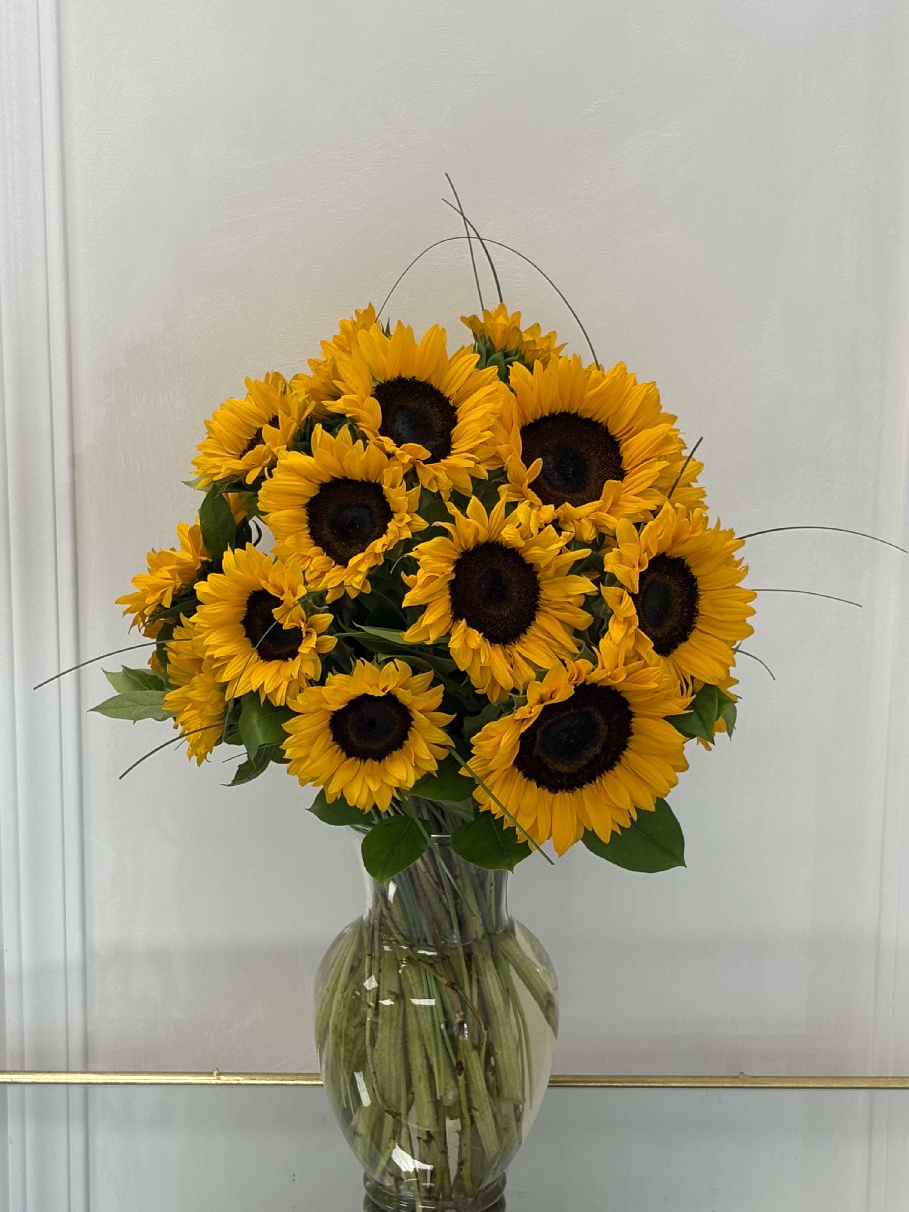 Summer Sunshine By VLM Flowers - A joyful burst of sunshine, this bouquet of radiant sunflowers is artfully arranged in a clear glass vase, showcasing their golden petals and dark centers. Lush greenery adds contrast, while the tall, graceful stems create a fresh-from-the-garden elegance. Perfect for brightening any room and lifting the spirit.