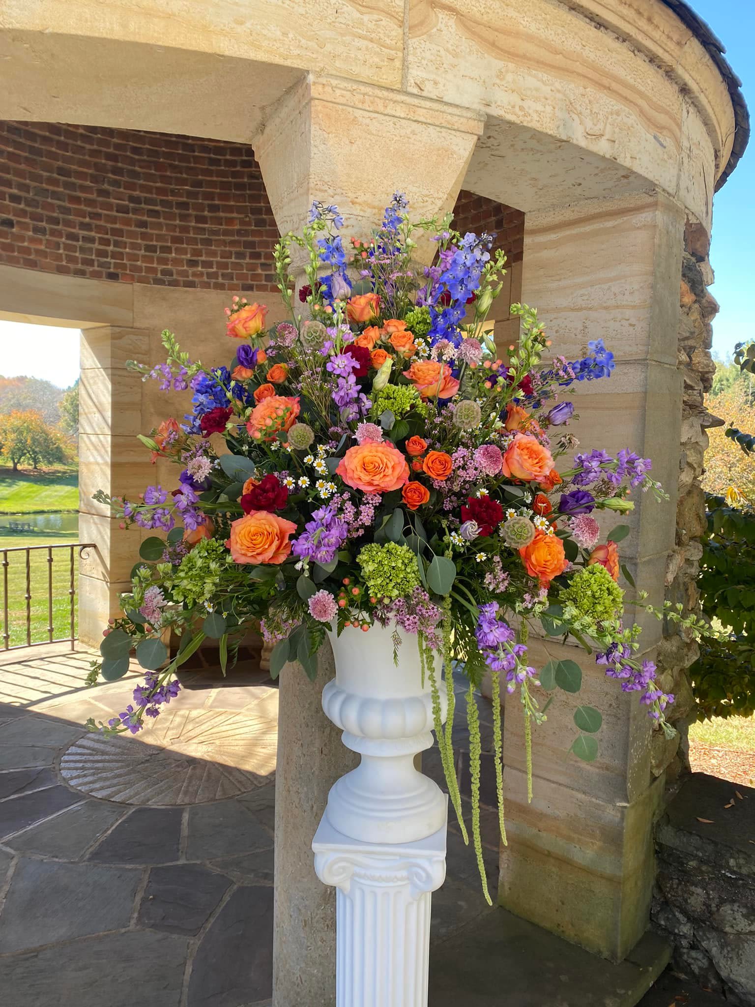 Glorious Garden - This glorious garden urn has blue delphinium, free spirit roses, lavender stock, green mini hydrangea, orange spray roses, carnations, monte casino and other fillers.