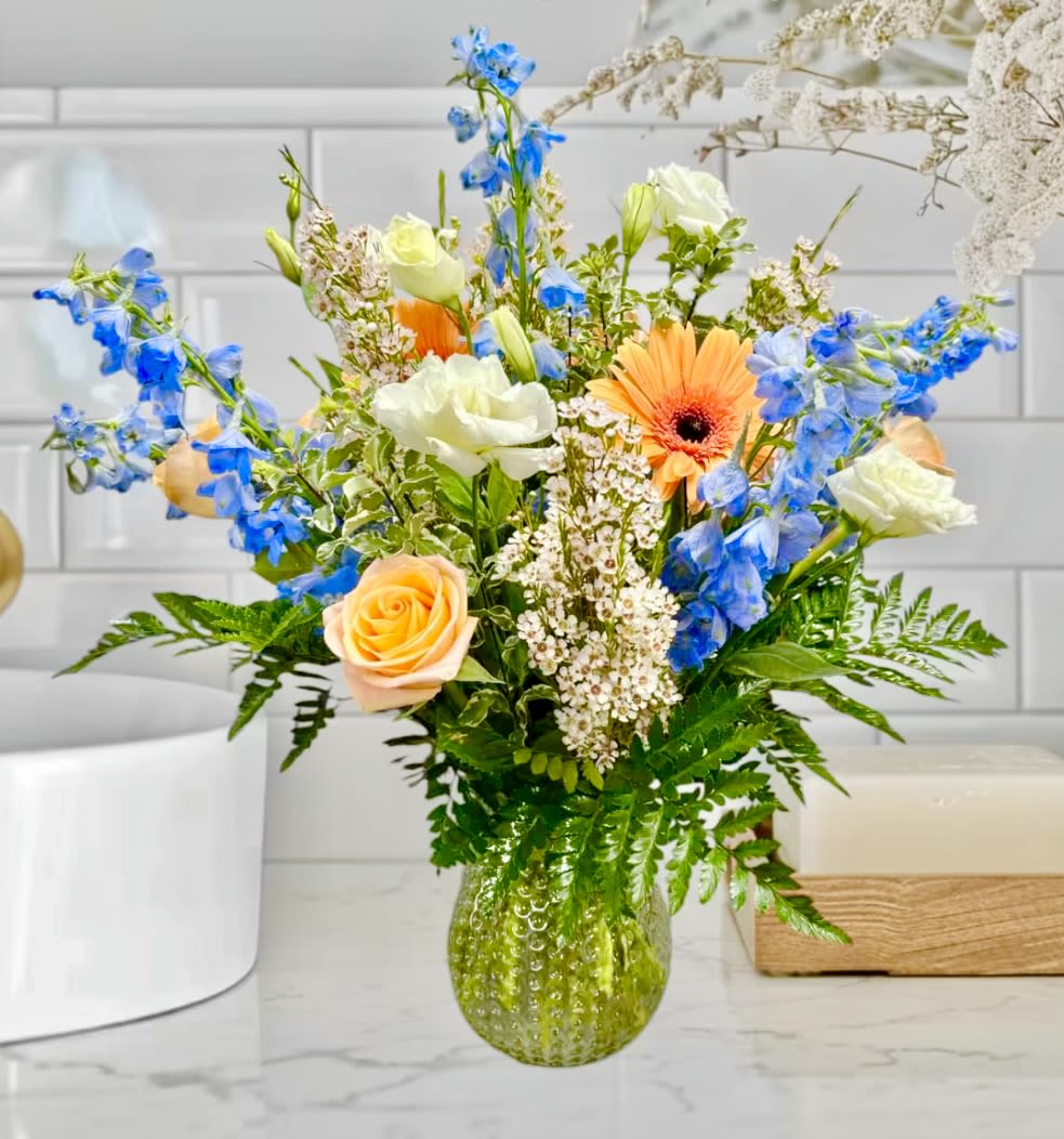 Just peachy! - This stunning vase features a delightful combination of orange gerberas and roses, blue delphinium, white lisianthus and white wax flower, creating a truly just peachy arrangement. (Substitutions may have to be made depending on color and flower availability. We will design the arrangement with the same money value but may have to make substitutions). 