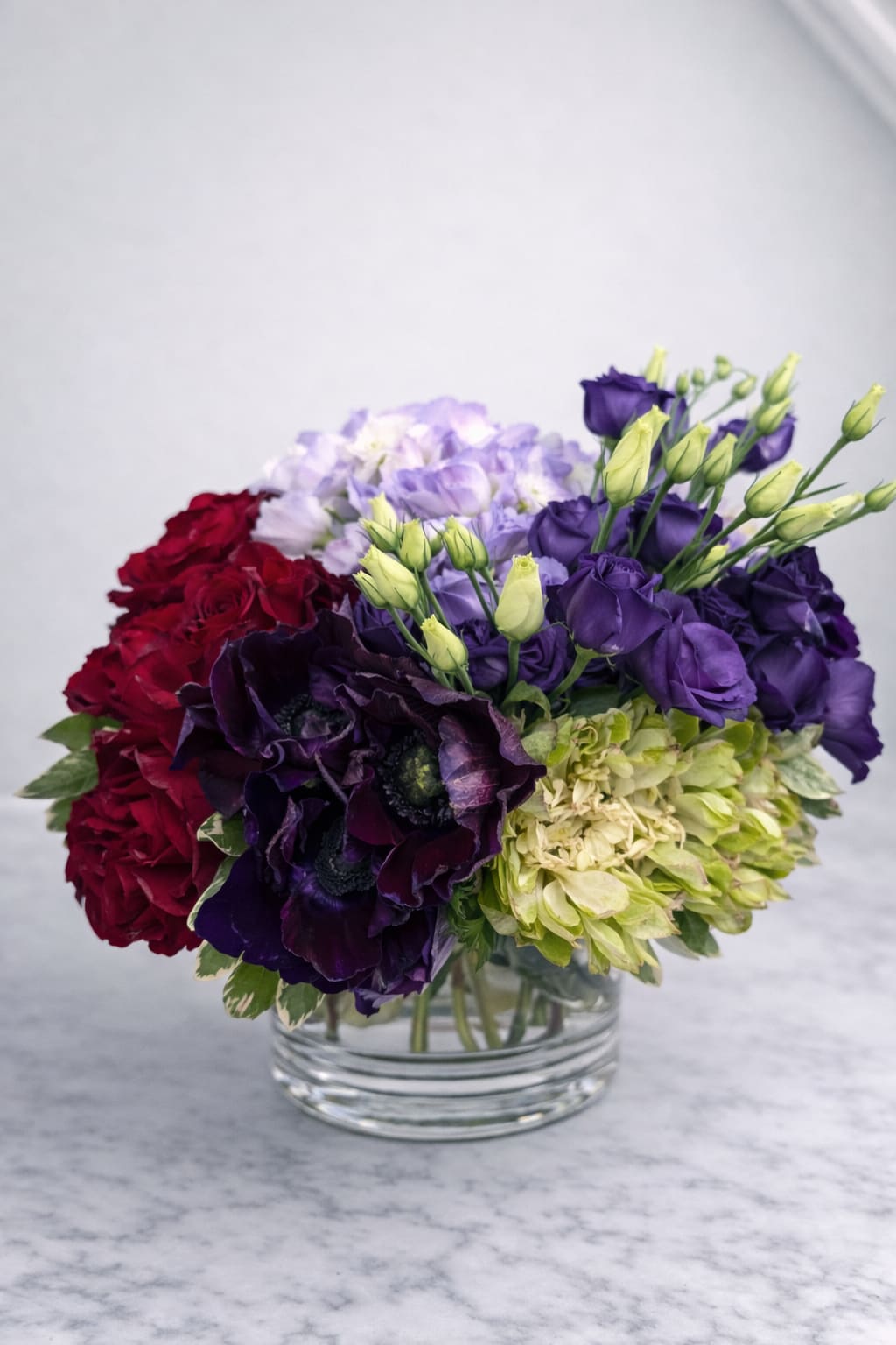 Twofold - Twofold is an exploration of contrast and harmony — a floral composition that reveals two distinct expressions within a single arrangement.  One side leans bold and passionate, layered with rich reds, deep plums, and dramatic textures. The other softens into loyalty and grace, with luminous greens, pale lavender, and gentle movement. Together, they create a quiet tension — a conversation between intensity and endurance, romance and restraint.  Designed to be viewed from multiple angles, Twofold invites you to slow down, turn the vase, and discover its shifting story. No single perspective tells the whole truth — and that’s exactly the point.  Perfect for those drawn to artistry, symbolism, and flowers that feel deeply intentional rather than decorative.