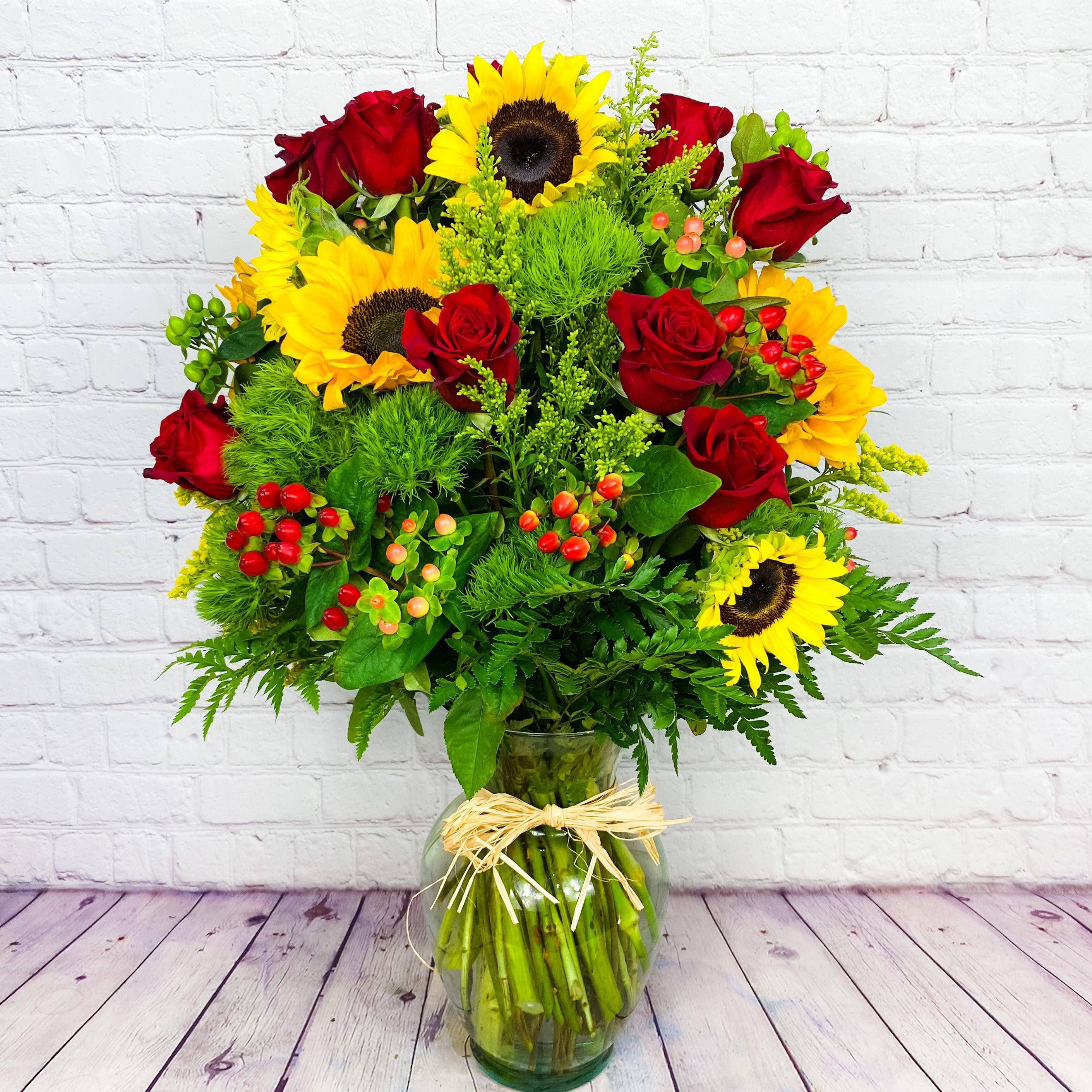 Rubies and Sunshine Bouquet - This captivating assortment of Fall's floral gems includes sunflowers,&nbsp;solidago, dianthus balls, hypericum berries and red roses...all artfully presented in a clear fluted vase. This Fall mixed flower bouquet is a fun gift to send during the Autumn season. Bring a touch of sunshine into your home or gift it to someone special with this eye-catching Sunflower bouquet. Experience the beauty and freshness of DGM Flowers today!   Includes:&nbsp; • Sunflowers • Red Roses&nbsp; • Hypericum •&nbsp;Solidago