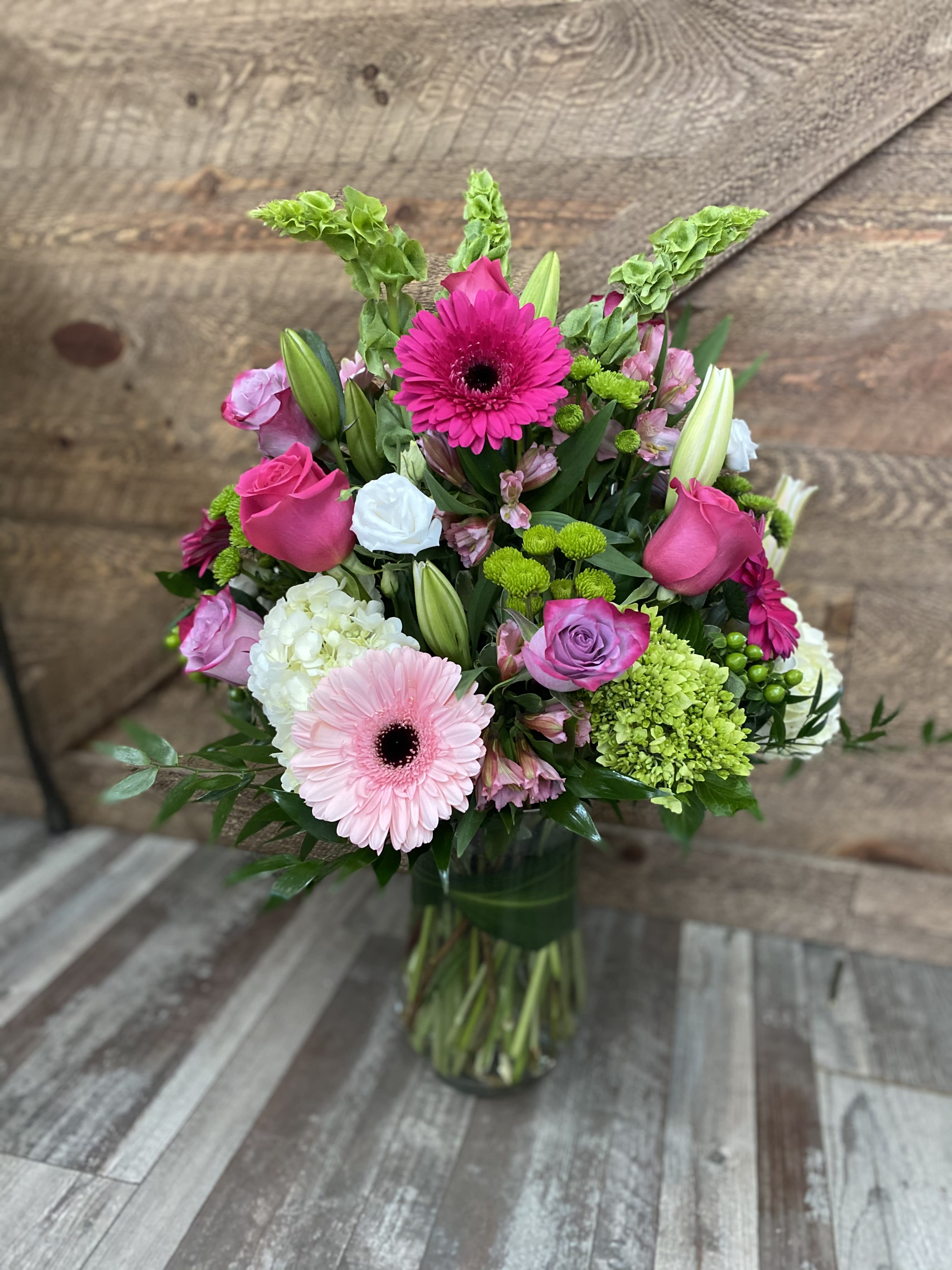 Sweetness - Gorgeous combination of flowers arranged taller style in a clear glass vase. Gerbera daisies, hydrangea, roses, alstromerias, mums, bells of Ireland and more. This design will be sure to impress. Version shown is the deluxe.