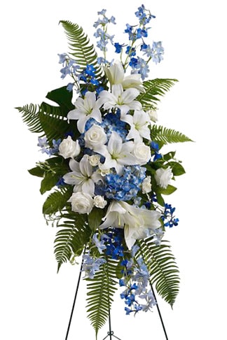 Ocean Breeze Standing Spray - Tranquil blue and white flowers reminiscent of a soft ocean breeze offer comfort. The standing spray presented on an easel includes fragrant white lilies and tropical greenery.
