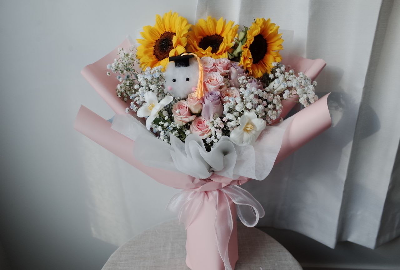 Golden Cheers Graduation Sunflowers Bouquet - For the graduate who deserves the world.   This joyful bouquet features radiant sunflowers—symbols of achievement and optimism—paired with delicate pink florals and a plush companions with graduation hat. It’s a sweet, celebratory gesture that says “I’m proud of you” in the most charming way.