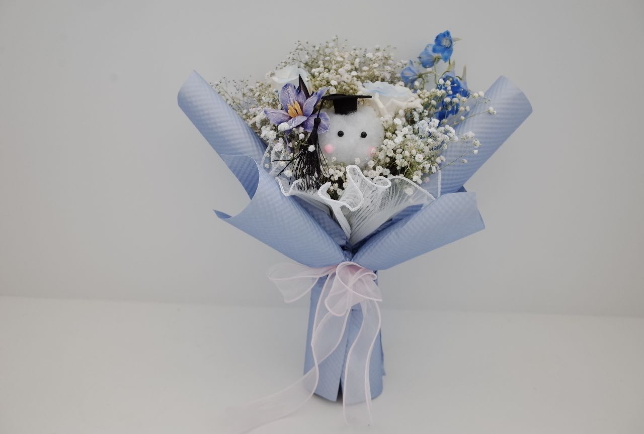 The Graduate’s Glow - For the star of the stage.   This bouquet blends elegance and playfulness: dainty white and purple flowers cradle a plush graduate in cap. Wrapped in pastel hues, it’s the perfect gift to honor achievement and send warm wishes for the future.