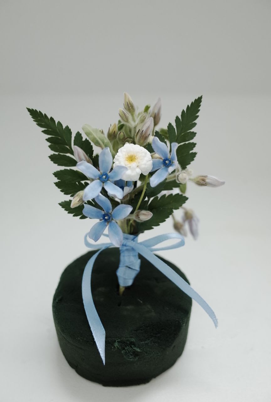 Blue Serenade Boutonniere - Light as a breeze, timeless as love.   Delicate florals in sky blue and ivory are gathered with fern-like greens and finished with a satin ribbon. This boutonniere is ideal for grooms, groomsmen, or fathers of the bride, offering a soft, sophisticated accent.