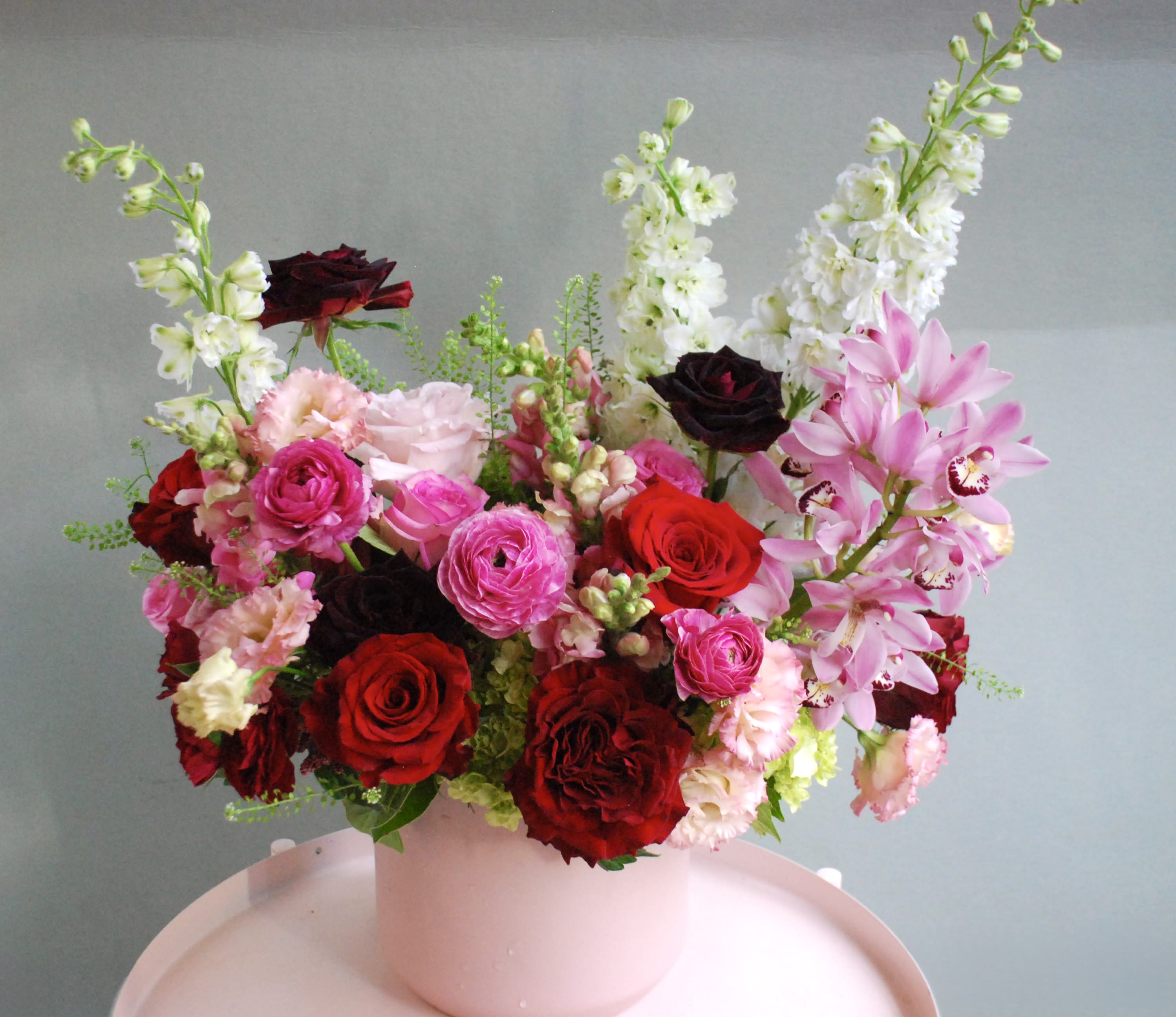 Never enough time - Garden-inspired arrangement blends deep reds  and hot pink roses with rich berry-toned blooms and playful seasonal accents. Textural layers of ranunculus, spray roses, and delicate greenery give this design a full, abundant feel. Styled in a white ribbed ceramic vase, it’s romantic, feminine, and effortlessly elegant—ideal for birthdays, celebrations, or just because. It will make you fall in love 