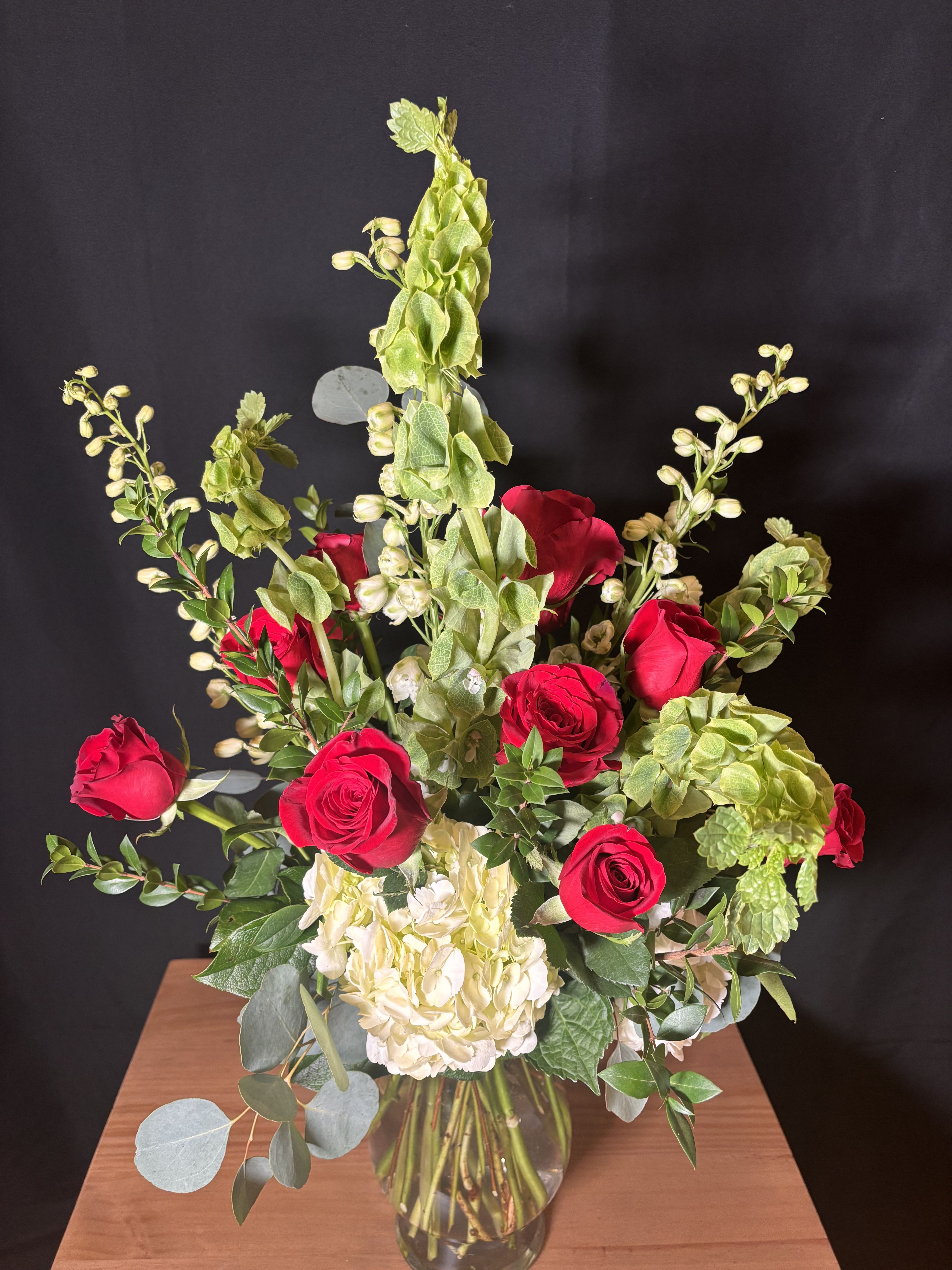 Crimson Garden Kiss - A timeless expression of love with a fresh, modern edge. This bouquet features a classic dozen velvety red roses nestled among soft hydrangea, sculptural Bells of Ireland, and layers of lush seasonal greens. Fragrant eucalyptus adds movement and a cool, aromatic finish, balancing the richness of the roses with natural elegance. Romantic, refined, and effortlessly beautiful—perfect for grand gestures or meaningful moments.
