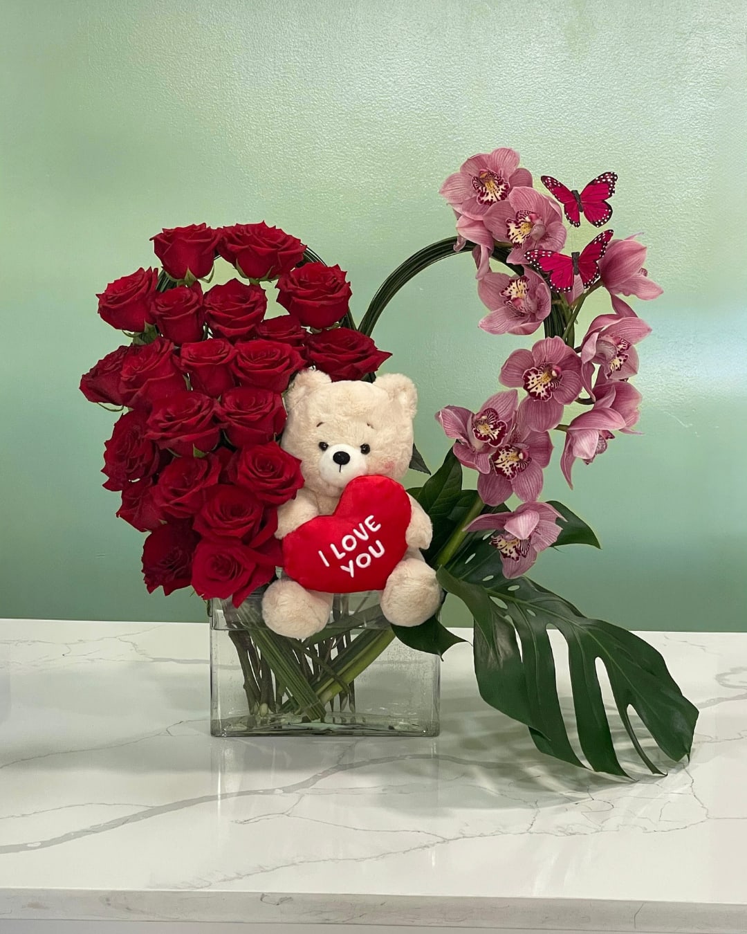 Lifetime of Love  - This marvelous arrangement has a cymbidium orchid, brilliant red roses, a beautiful monstera leaf, with a soft teddy bear sitting at the center and adorned with two butterflies.