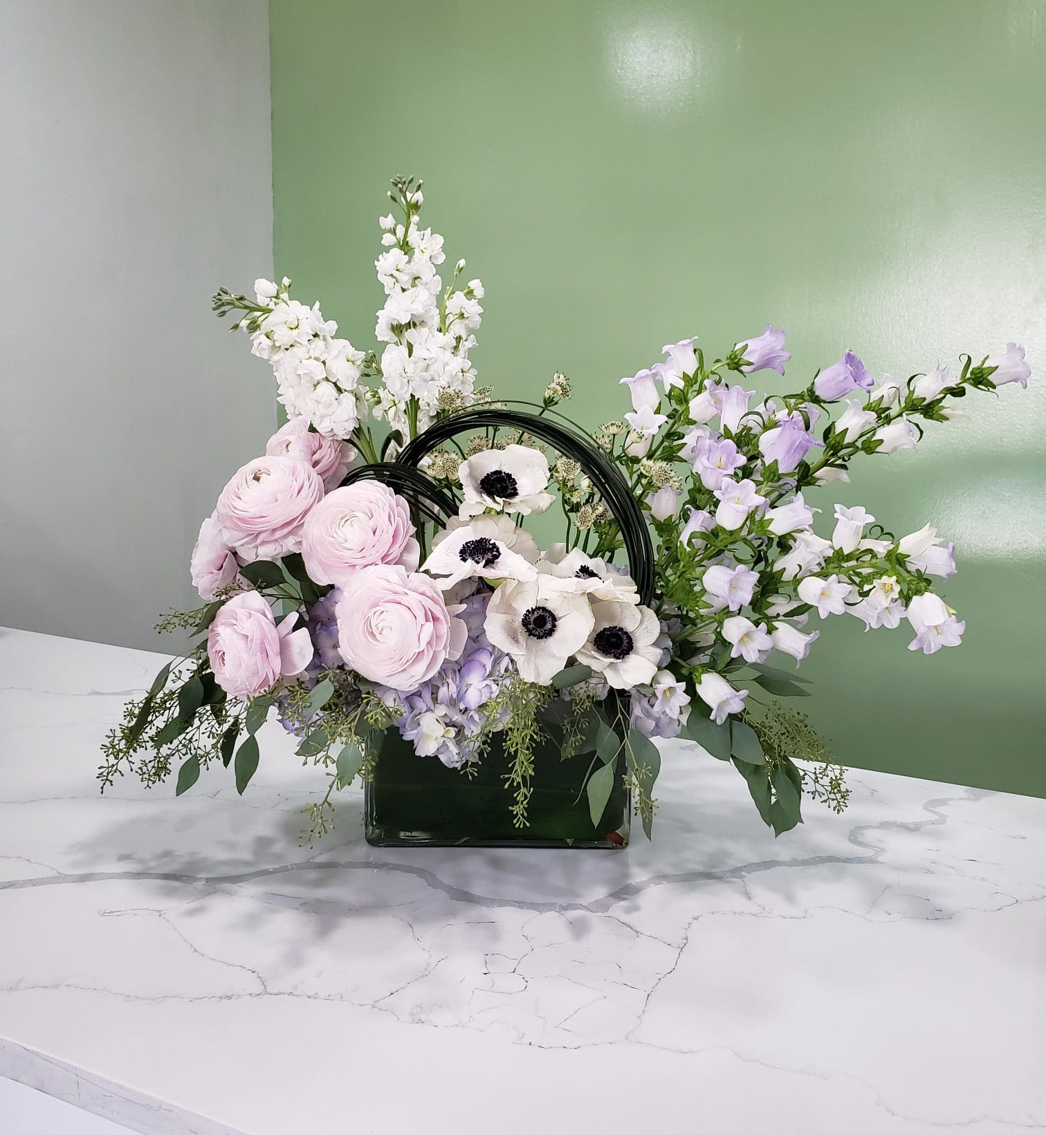 Once Upon a Time - This most dream-like arrangement has anemones, light purple hydrangeas, white stock, light pink ranunculus, light lavender campanula flowers, greens and white raster in a clear, rectangular, glass vase.