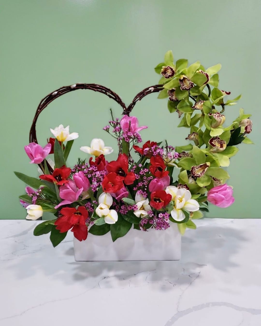 Blooming Affection - This magnificent arrangement has pink, white, red tulips, pink wax, green cymbidium orchid, and pussywillow branches in the shape of a heart in a white ceramic rectangular vase. 